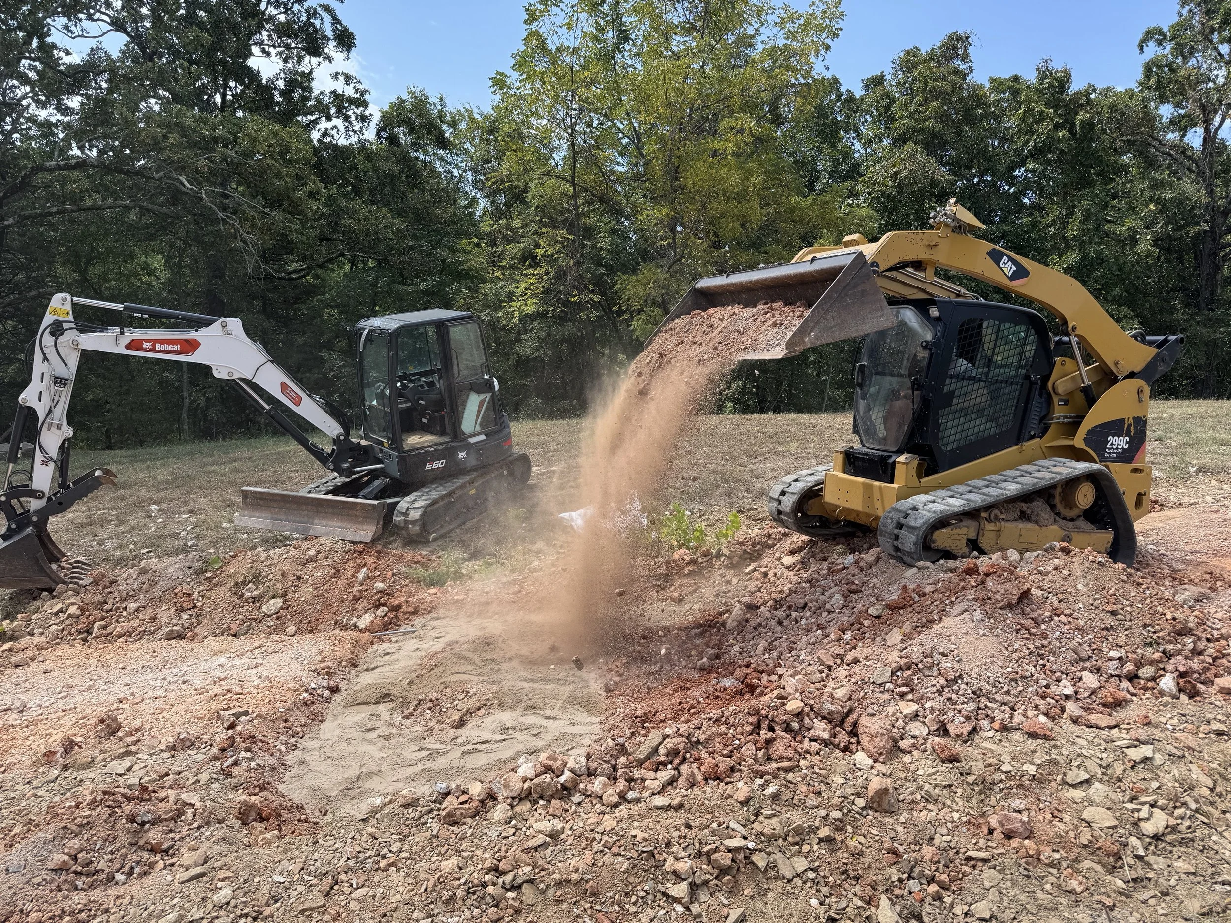 Construction site with two excavators moving dirt, surrounded by trees under a blue sky.
