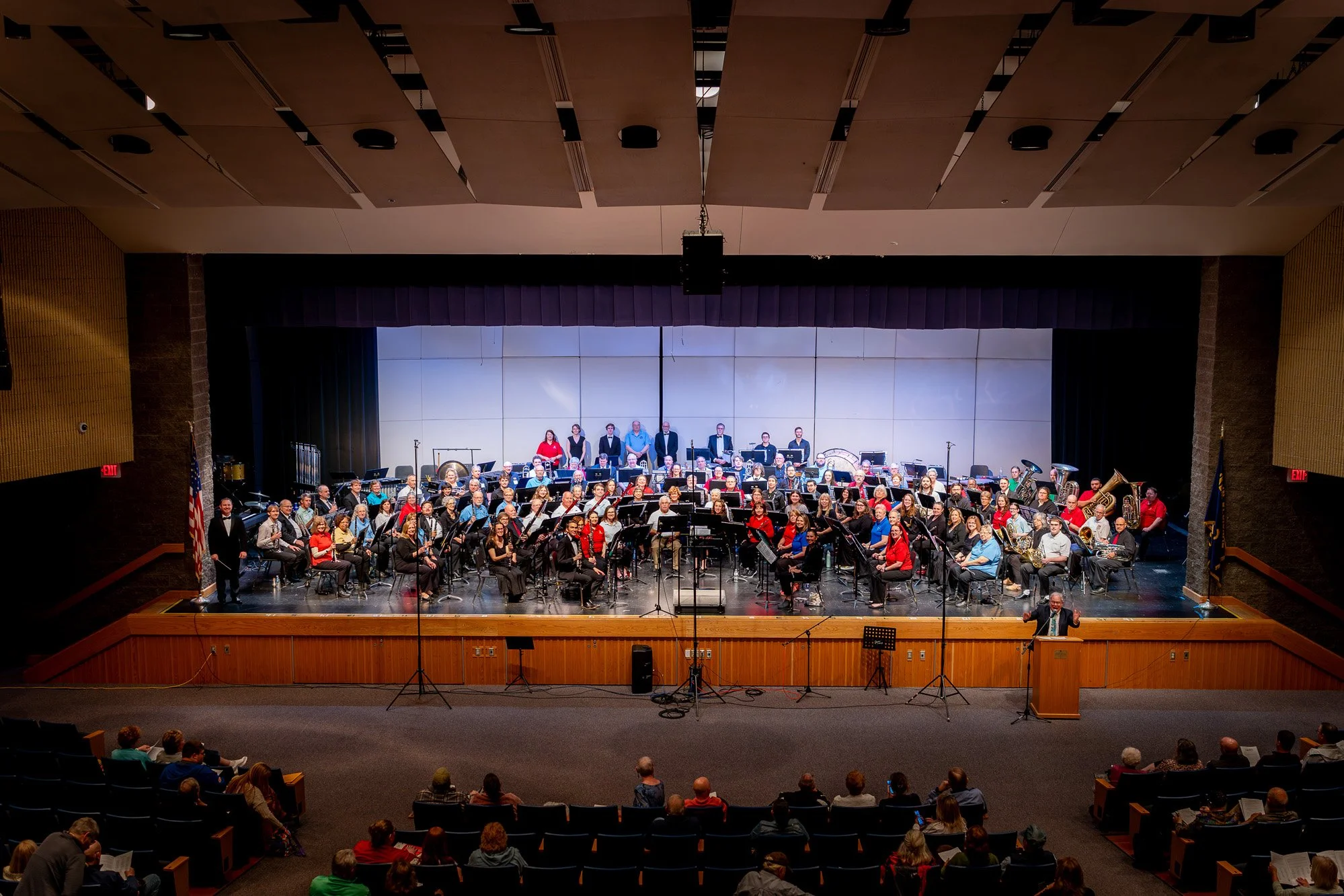 Full concert band seated on a high school auditorium stage with conductor at left and audience visible in the foreground.
