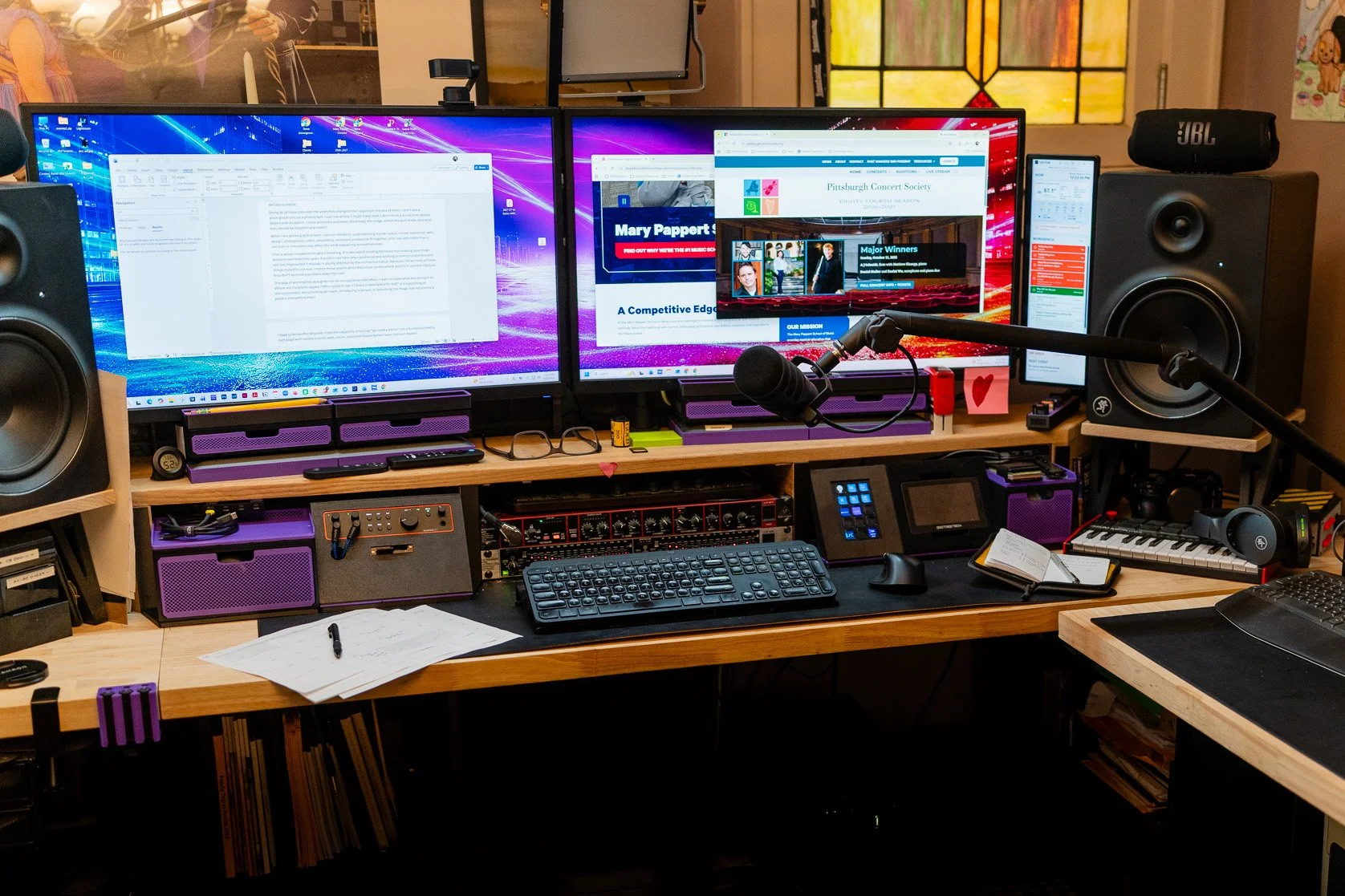 A busy multimedia workspace with a blog draft on one monitor and Pittsburgh Concert Society and Duquesne web pages on another, surrounded by a microphone, audio equipment, keyboards, and production controls.