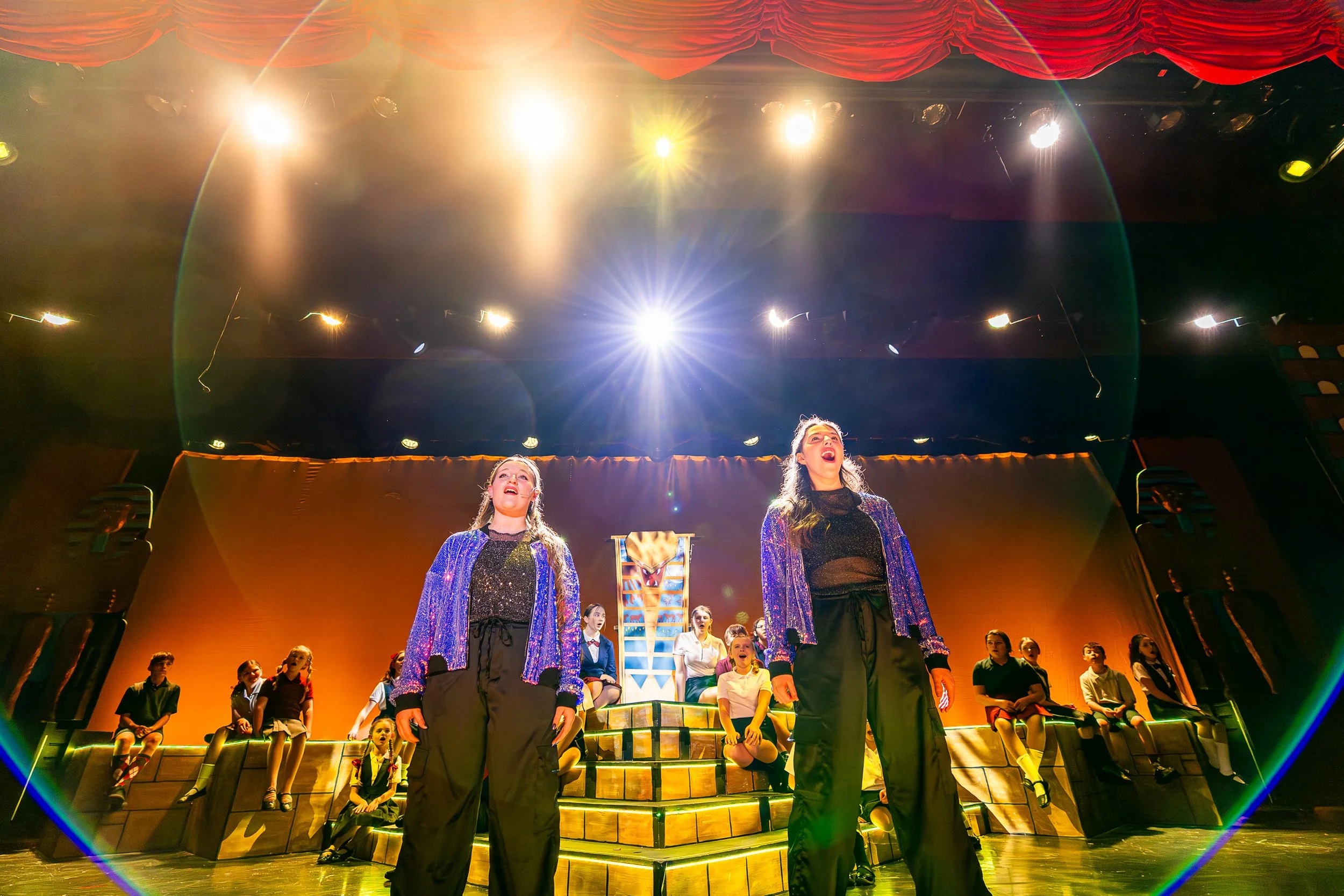 Two people singing on stage in front of a choir of children. Stage lights line the top of the photo, creating circular lens flares around the singers.