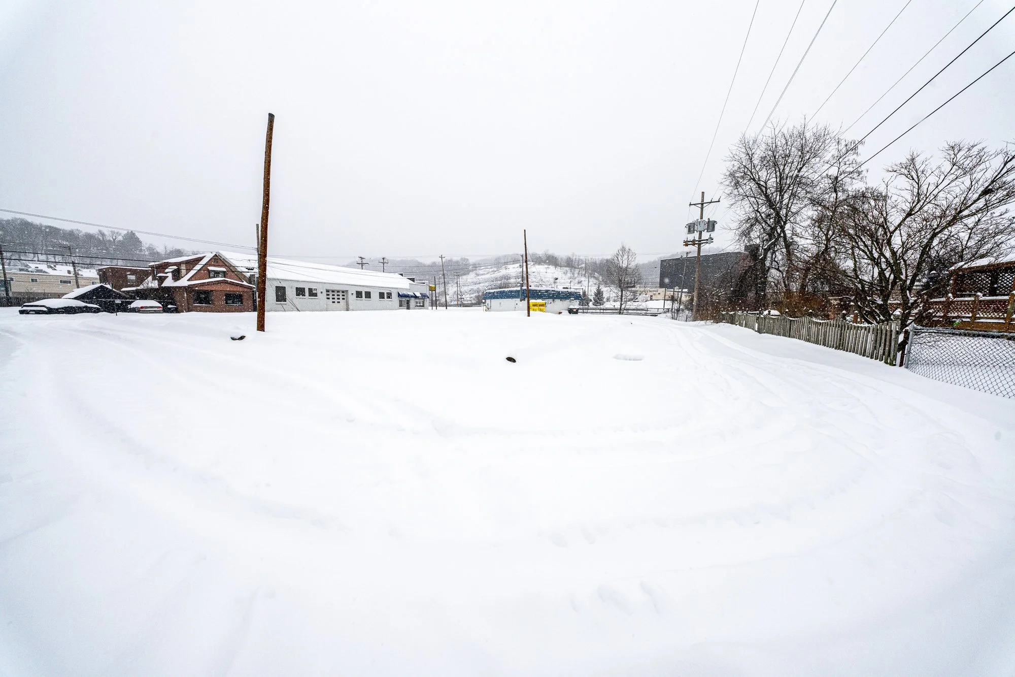 Snow-covered empty parking lot during active snowfall, with low buildings, utility poles, and distant hills barely visible through the winter haze.