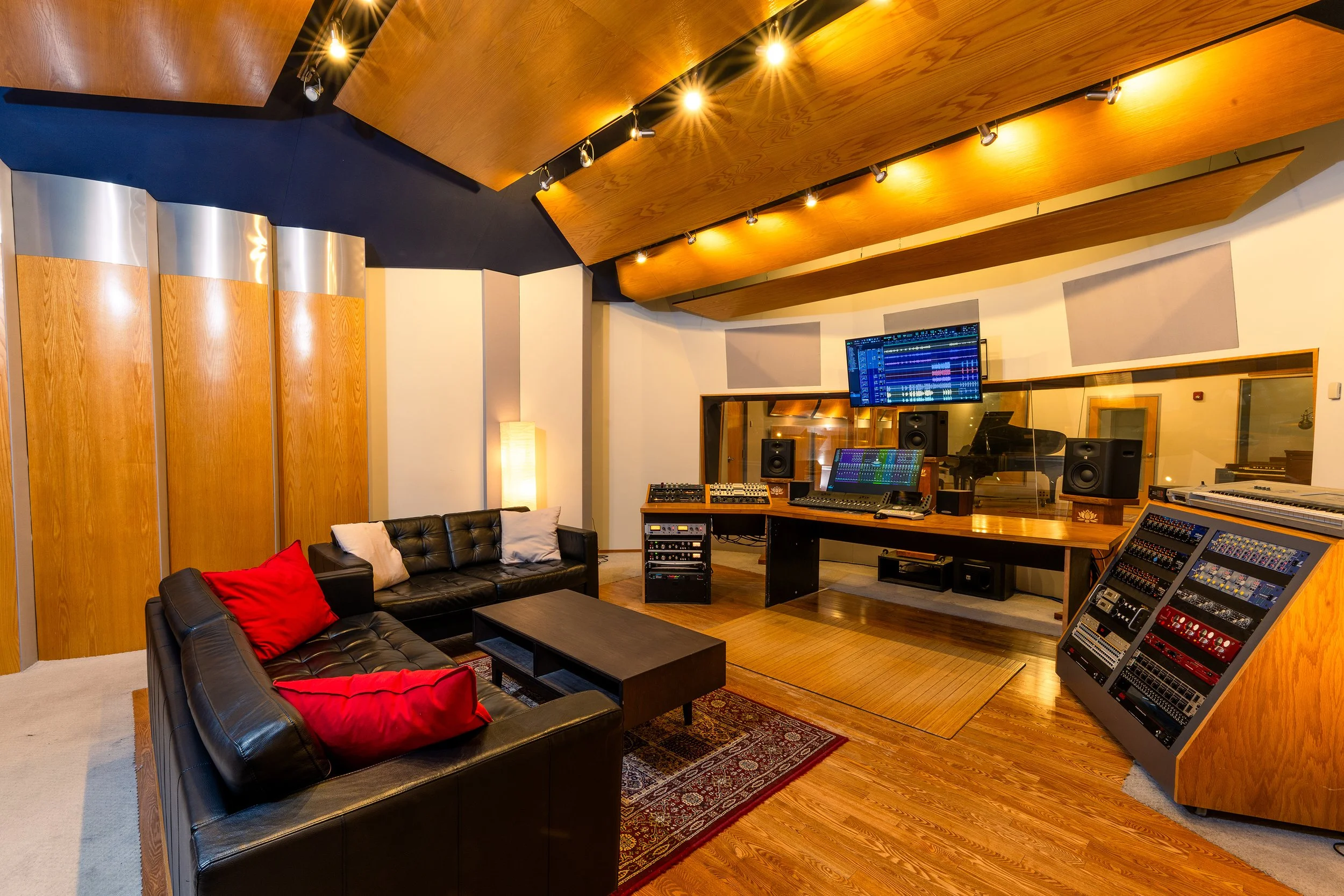 Recording studio control room with leather couches, red accent pillows, hardwood floors, a large mixing console, studio monitors, and a computer display showing audio tracks beneath curved wooden acoustic panels.