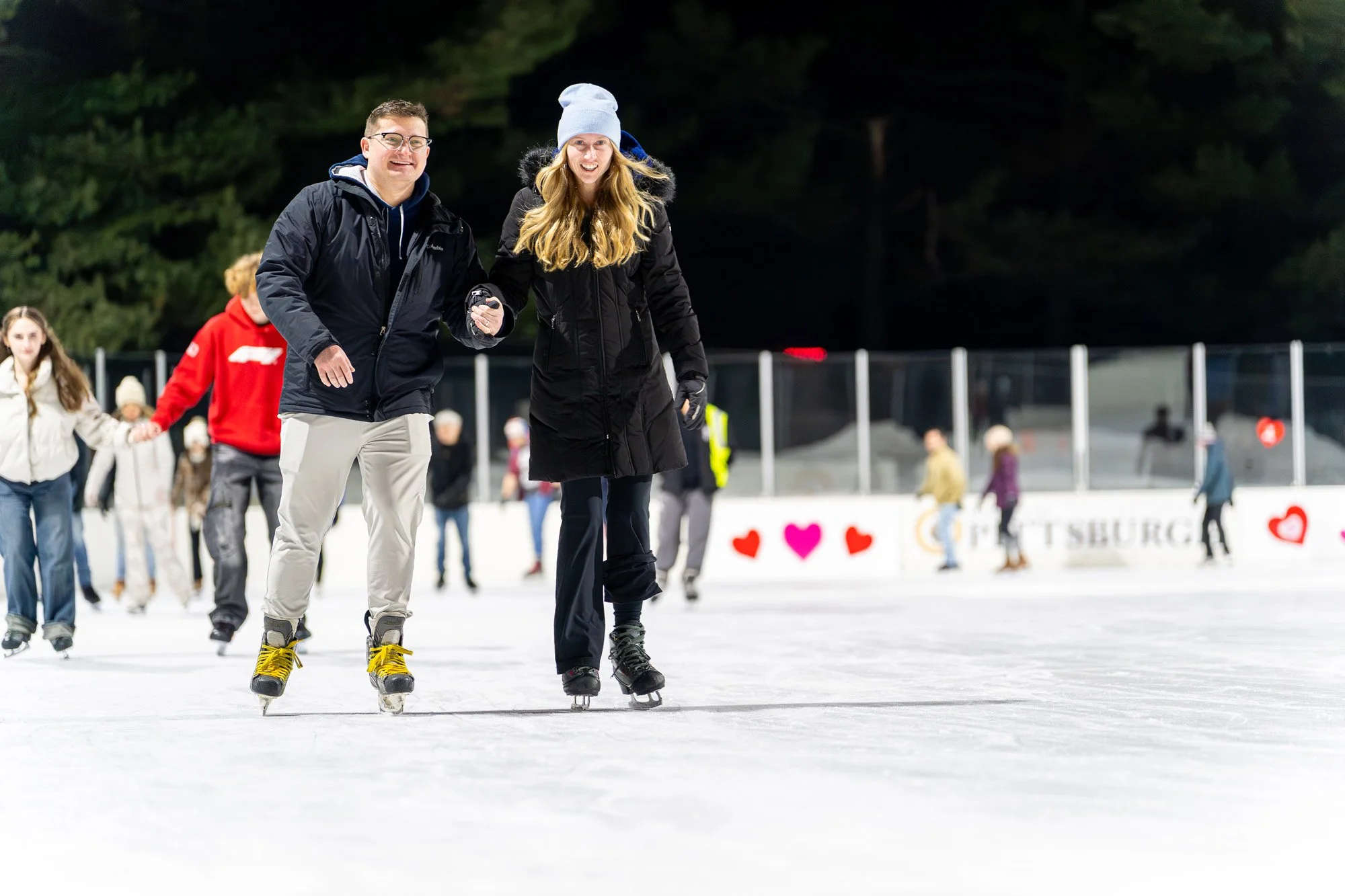 Couple ice skating hand in hand at an outdoor rink during a winter evening event, smiling as other skaters glide in the background with heart decorations along the boards.