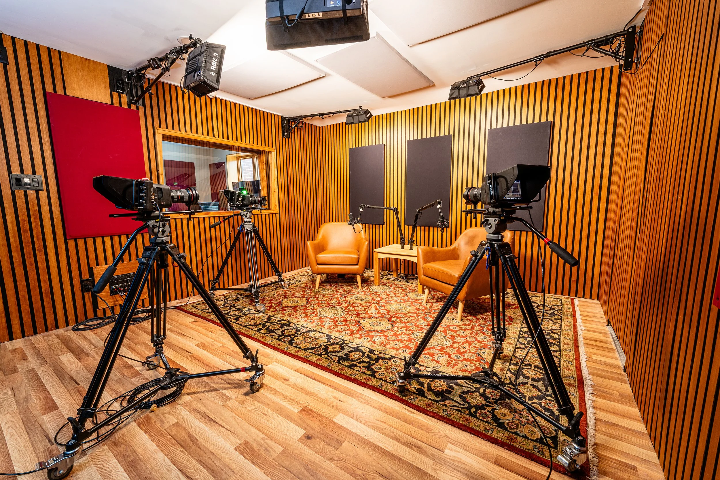 Video recording studio with three cameras on tripods facing a pair of leather chairs and microphones, set on a patterned rug beneath studio lights and wood acoustic wall panels.