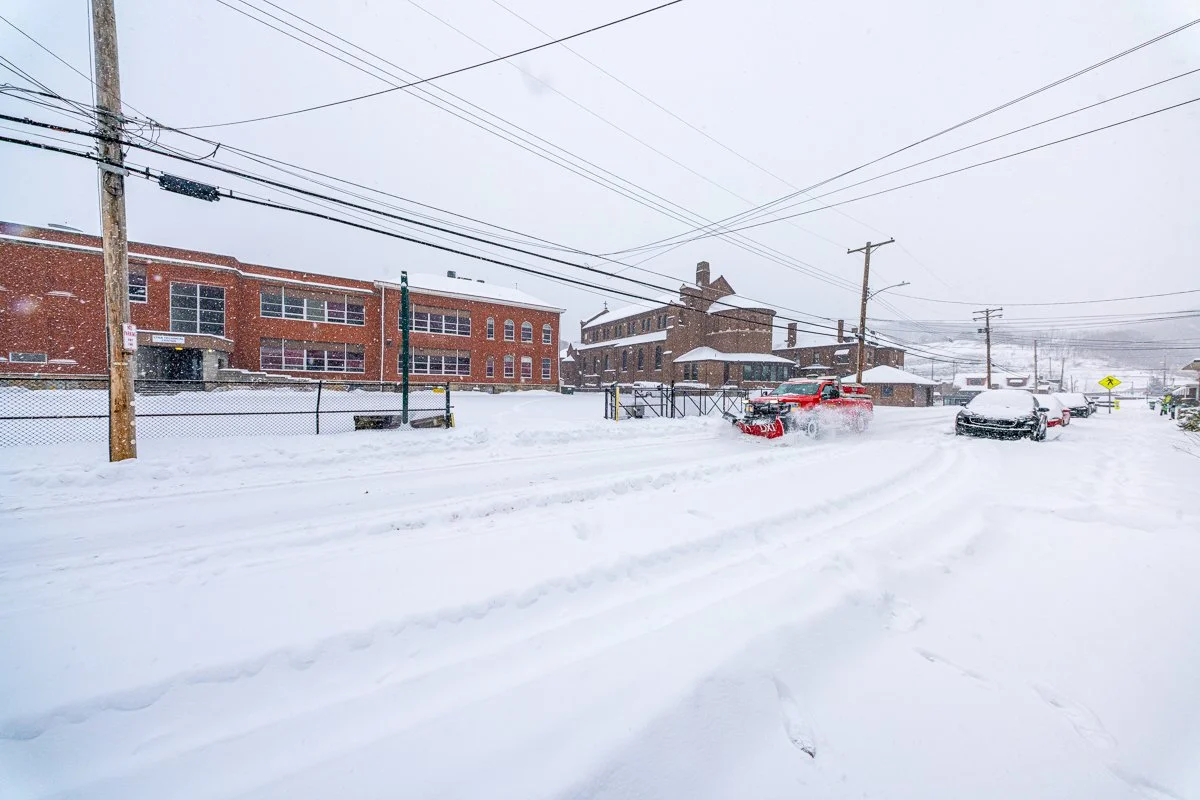Snowplow clears a residential street during active snowfall, passing brick buildings and parked cars while fresh tire tracks cut through deep snow.