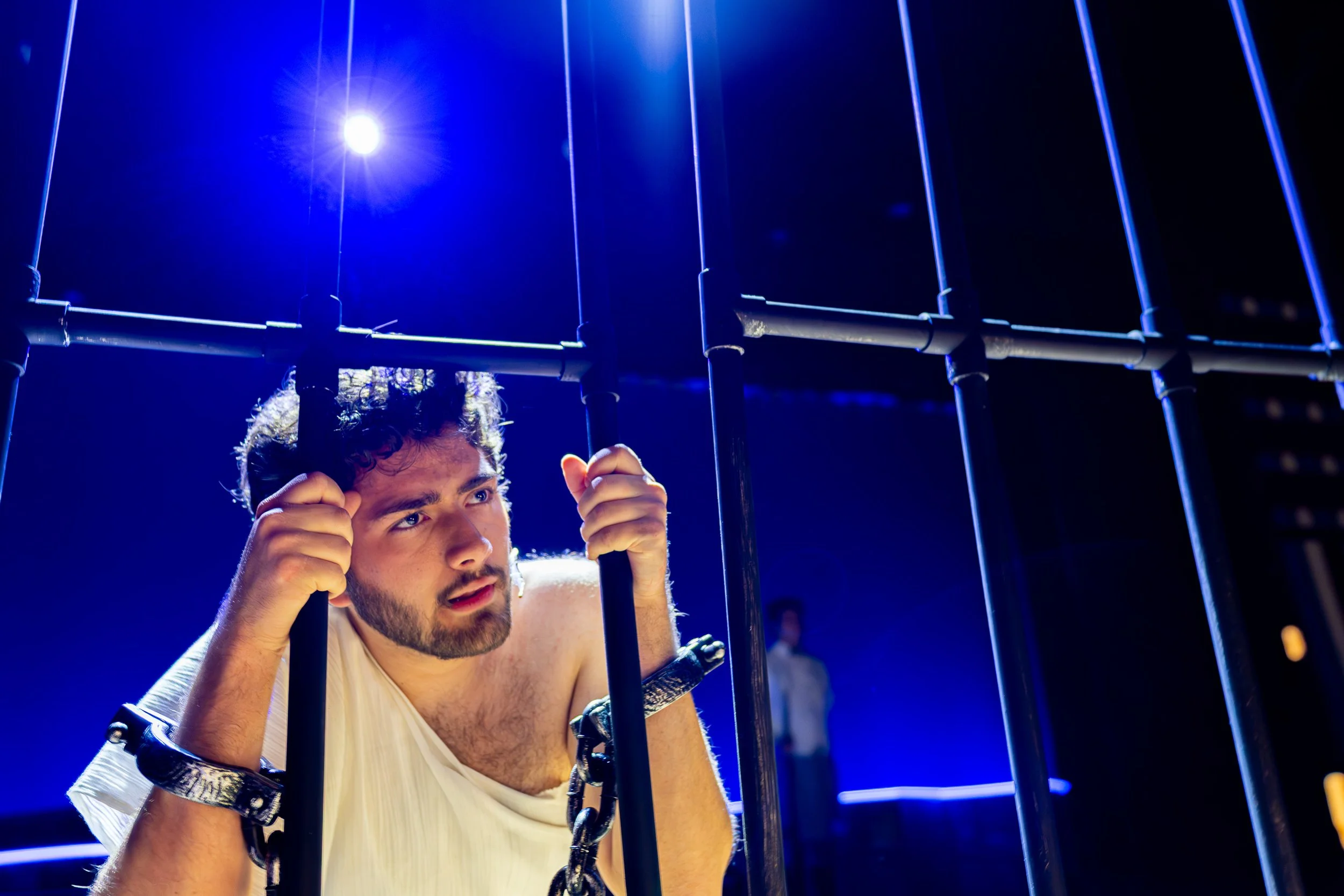 A person in handcuffs holds on to jail cell bars while singing on stage, lit from behind.