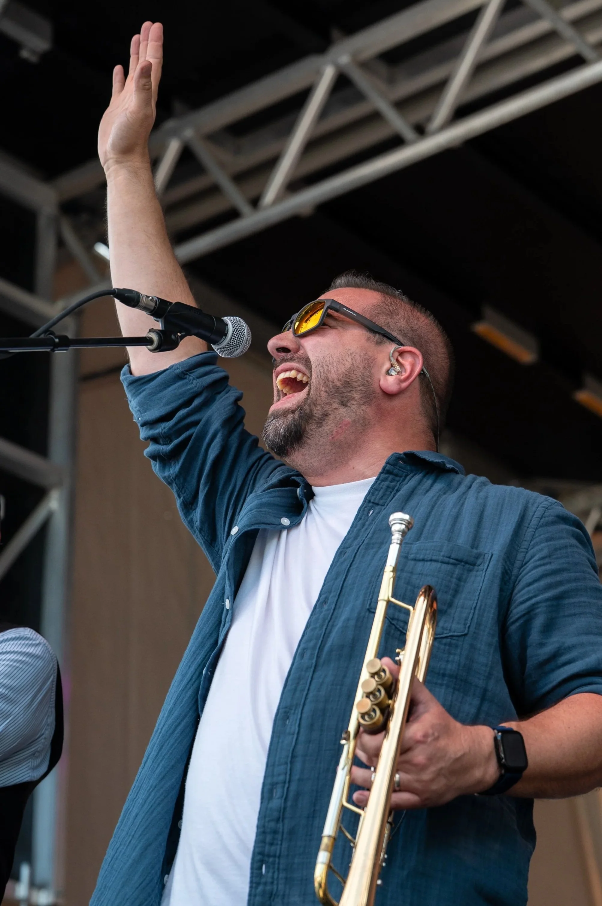 Person wearing sunglasses and holding a trumpet raises their hand in the air and sings in to a microphone.