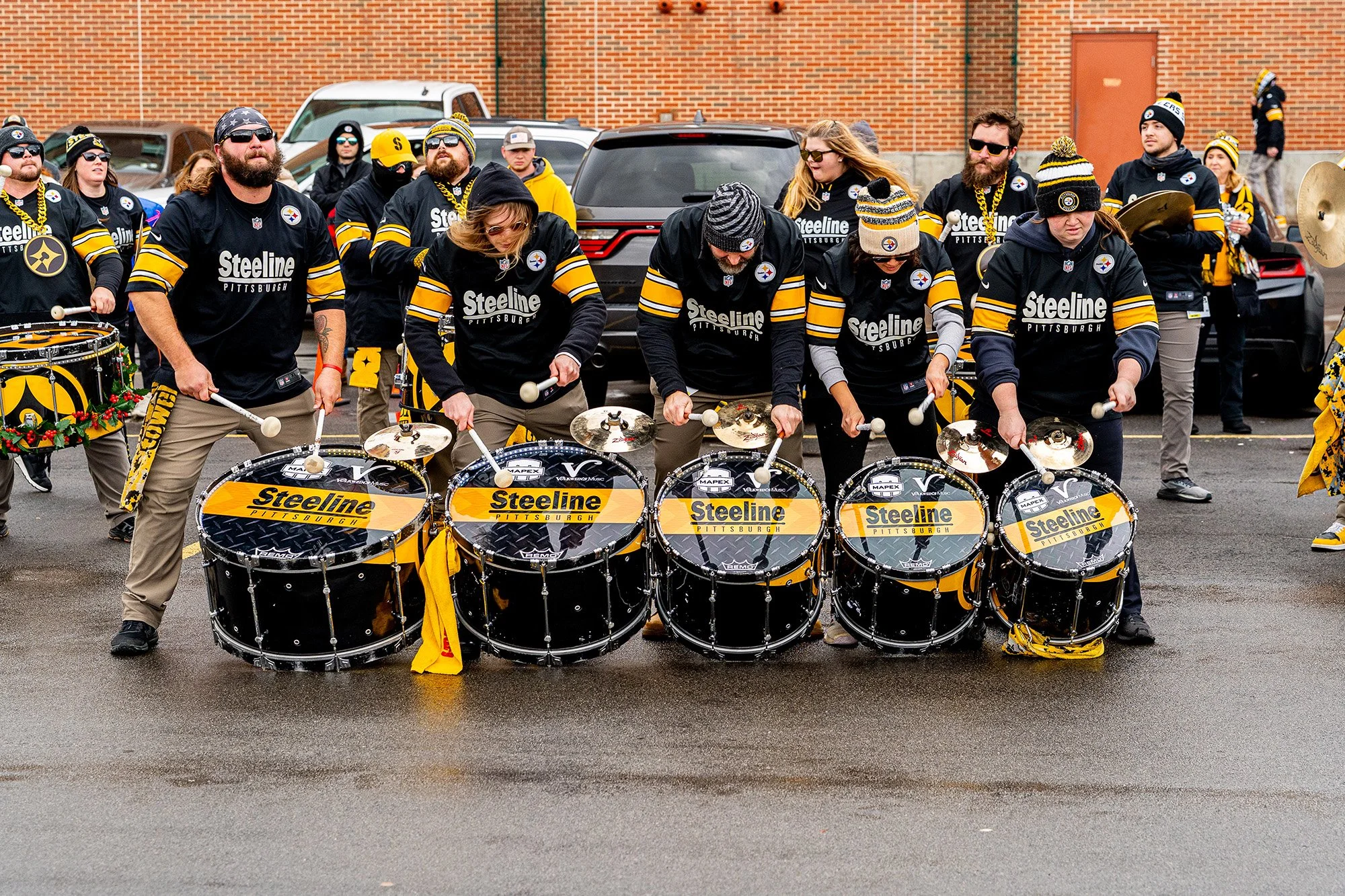 Drumline performers in black and gold Pittsburgh jerseys play bass drums in formation during an outdoor parade, with bundled spectators and parked cars behind them on a wet street.