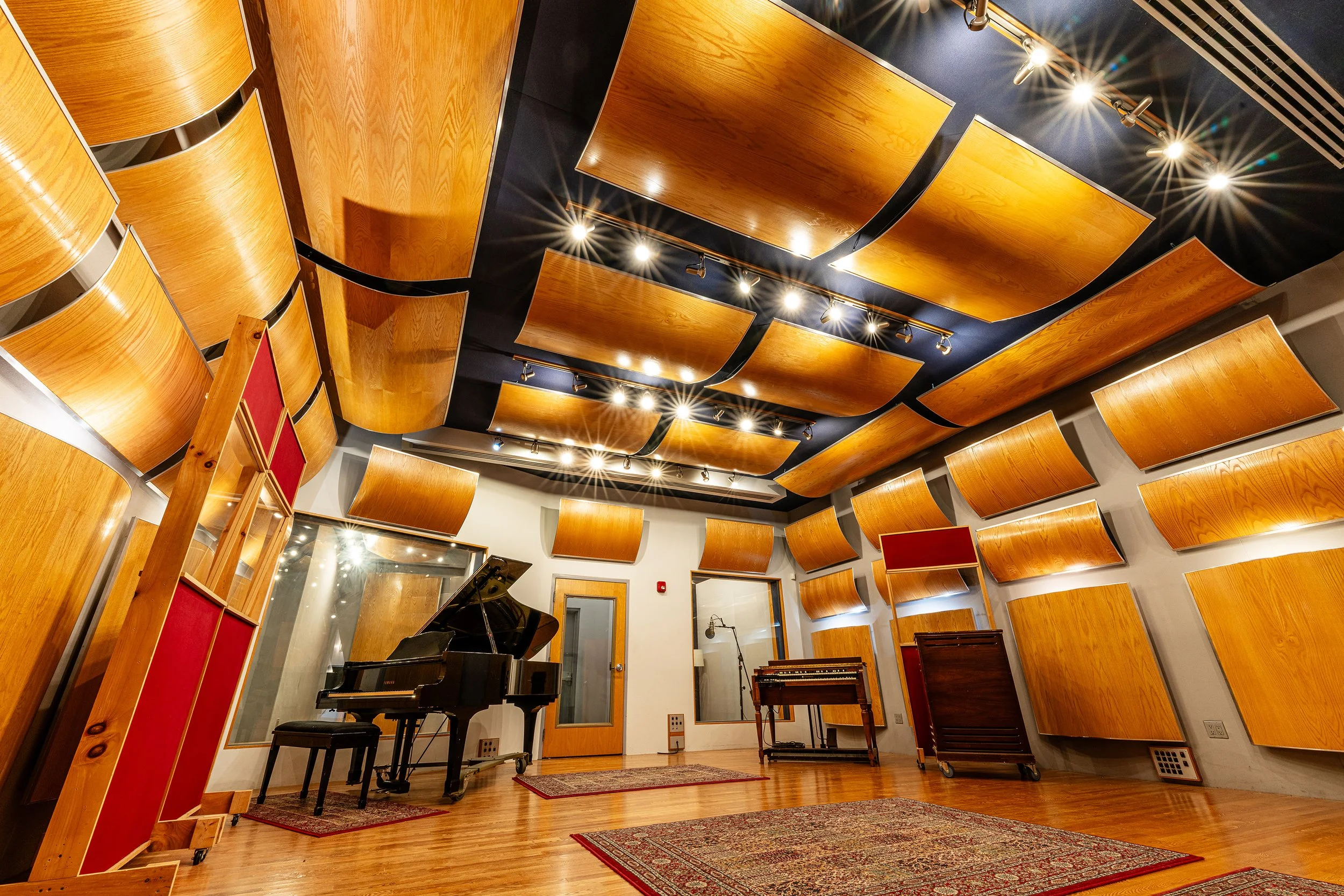 Recording studio interior with curved wooden acoustic panels on the walls and ceiling, a grand piano on a rug, and studio lighting illuminating the space.
