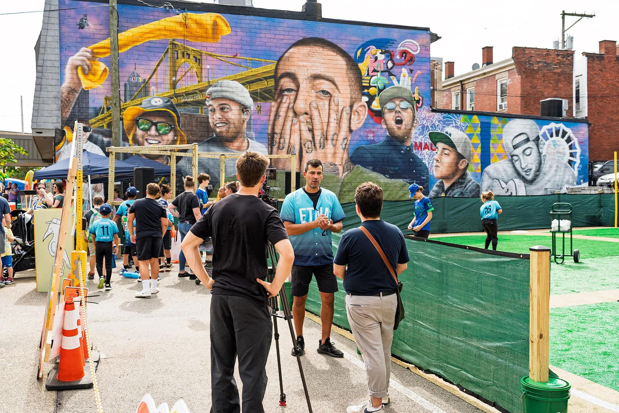 Person in a teal jersey speaks to two people beside a camera tripod at an outdoor community event, with children playing on turf and a colorful mural covering a brick building in the background.