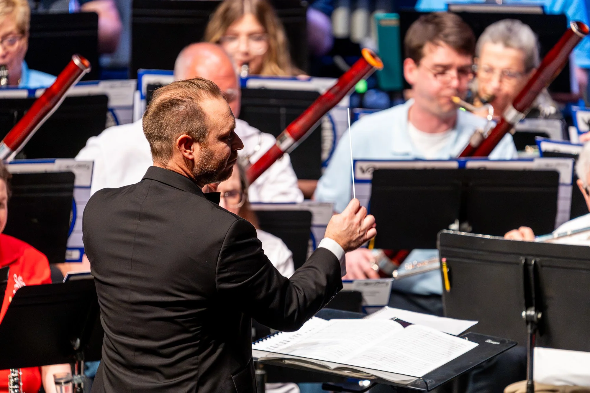 Conductor in black suit leading a concert band, holding a baton mid-gesture with woodwind players blurred in the background.