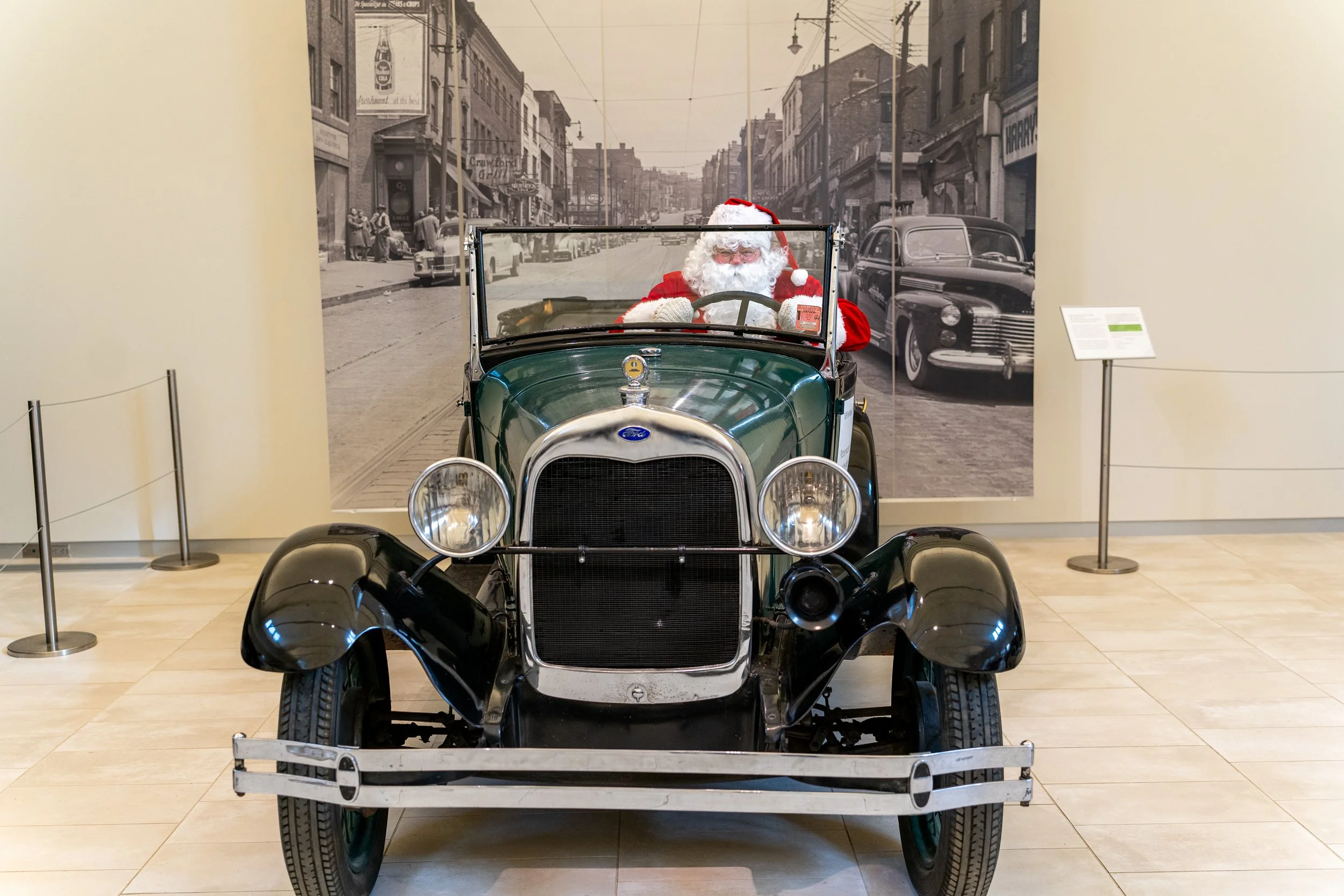 Santa Claus seated in a vintage green Ford Model A inside a museum, with a historic street mural behind the car.
