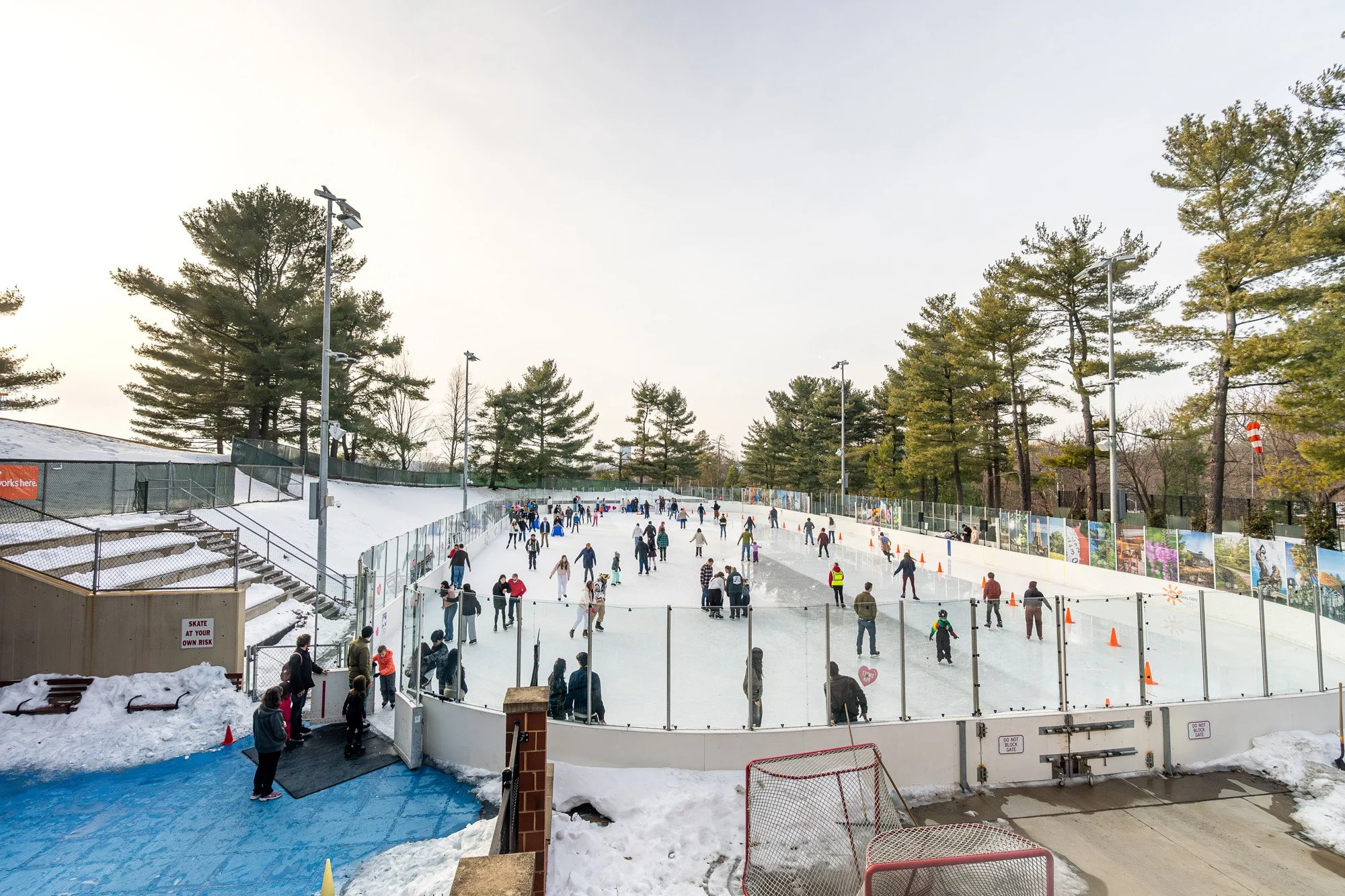 Wide view of a crowded outdoor ice rink with dozens of skaters circling the ice, surrounded by trees and glass boards, as spectators gather near the entrance on a winter afternoon.