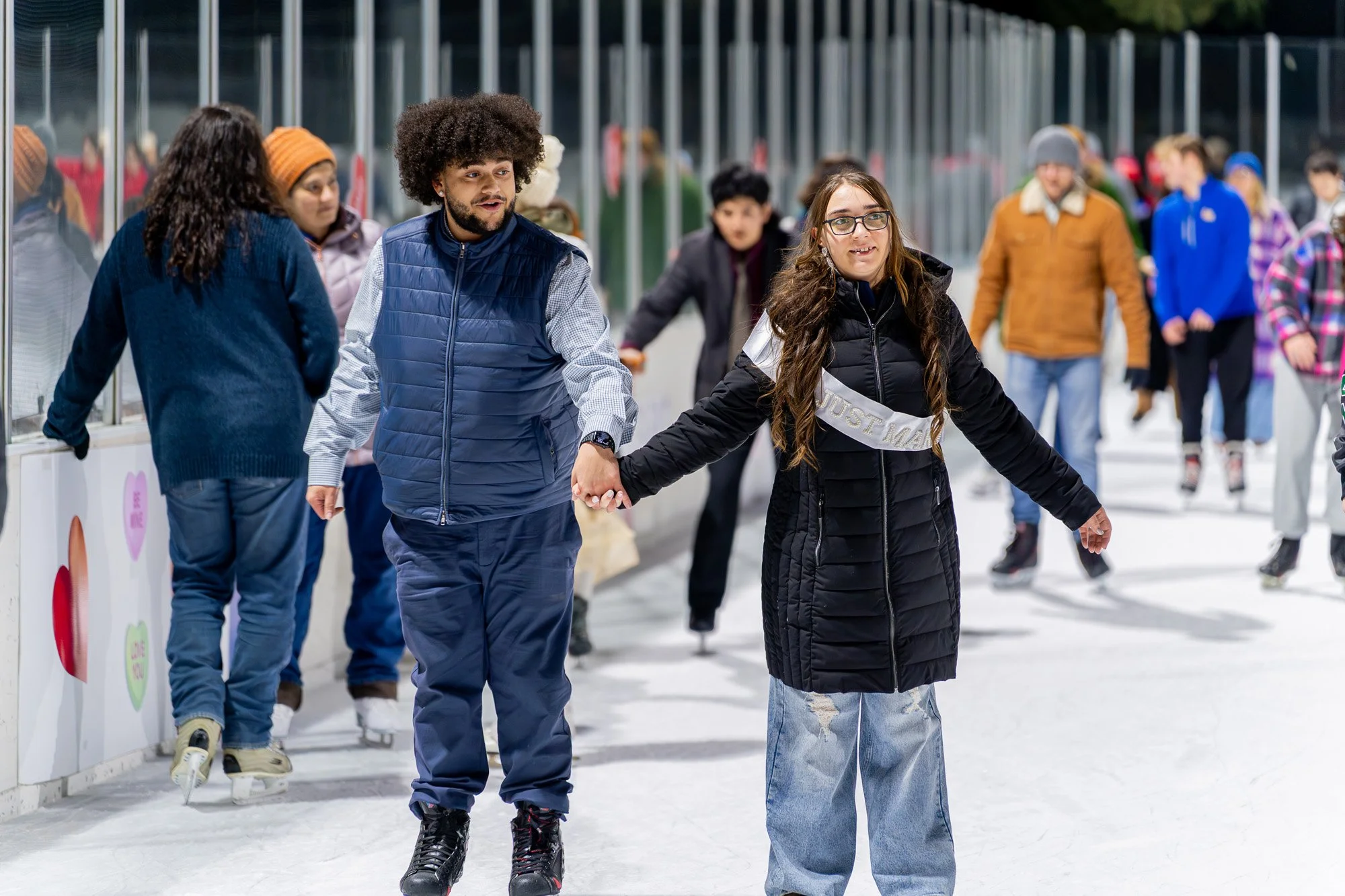 Couple ice skating hand in hand at an outdoor rink during a winter event, with the woman wearing a “Just Married” sash as other skaters glide past heart-decorated boards in the background.