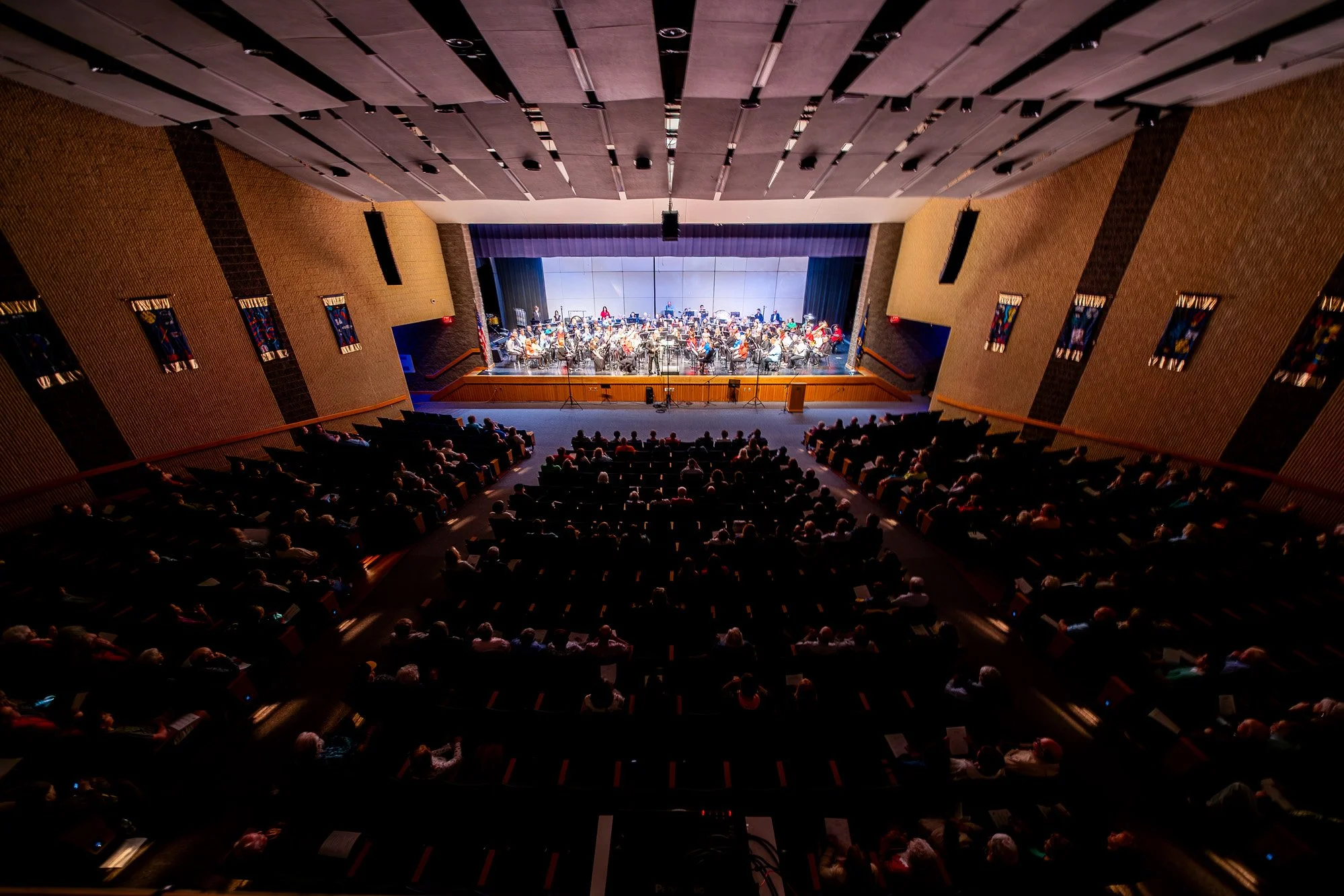 Wide view from the balcony of a large auditorium with a concert band performing on stage and audience seated below.