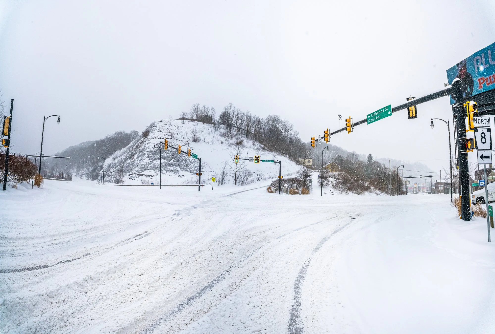 Snow-covered intersection with traffic lights and road signs, tire tracks cutting through fresh snow, and a rocky hillside in the background during active snowfall.