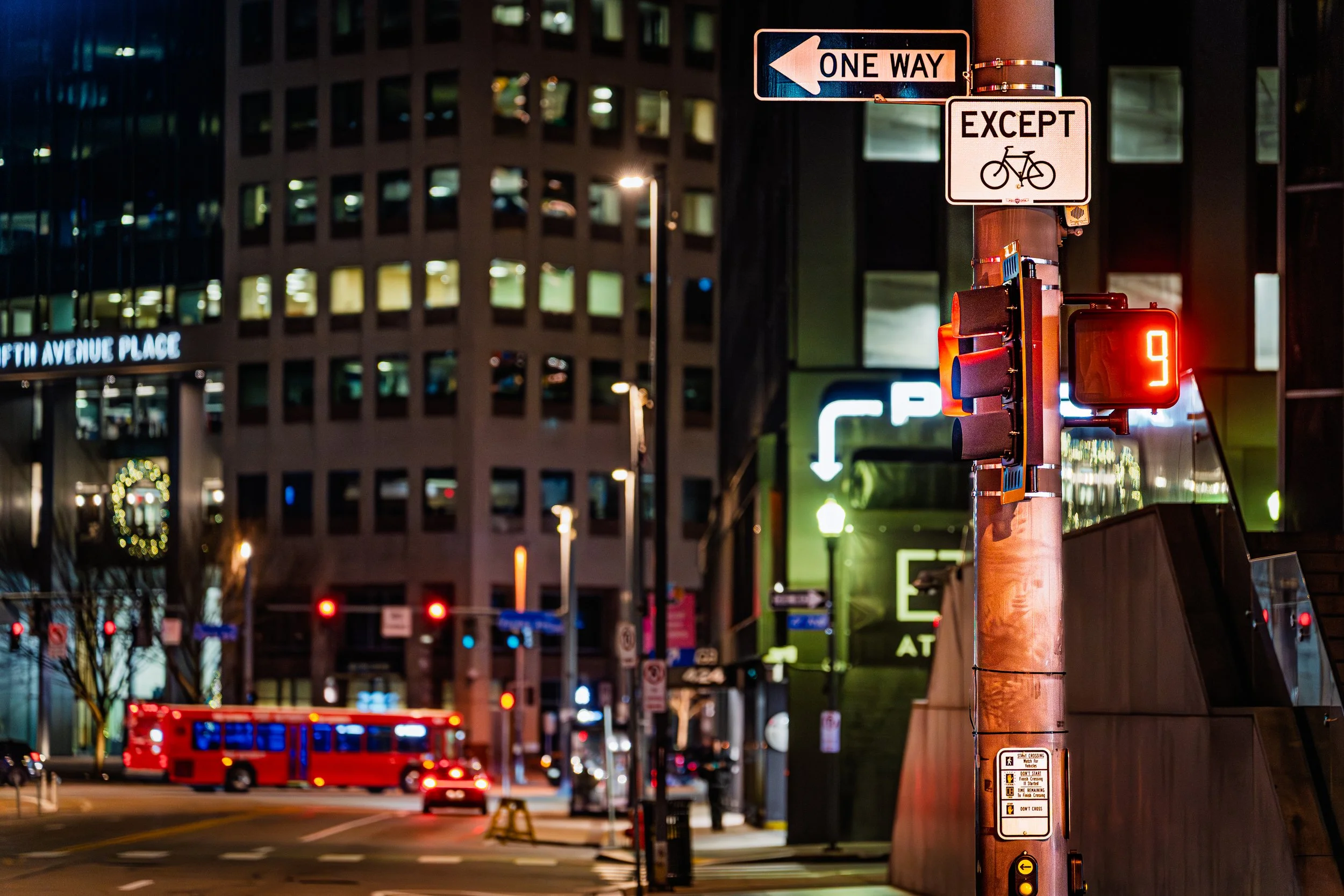 Nighttime downtown Pittsburgh intersection with a One Way sign and bicycle exception above a red pedestrian countdown signal showing nine seconds, illuminated buildings and a red city bus in the background.