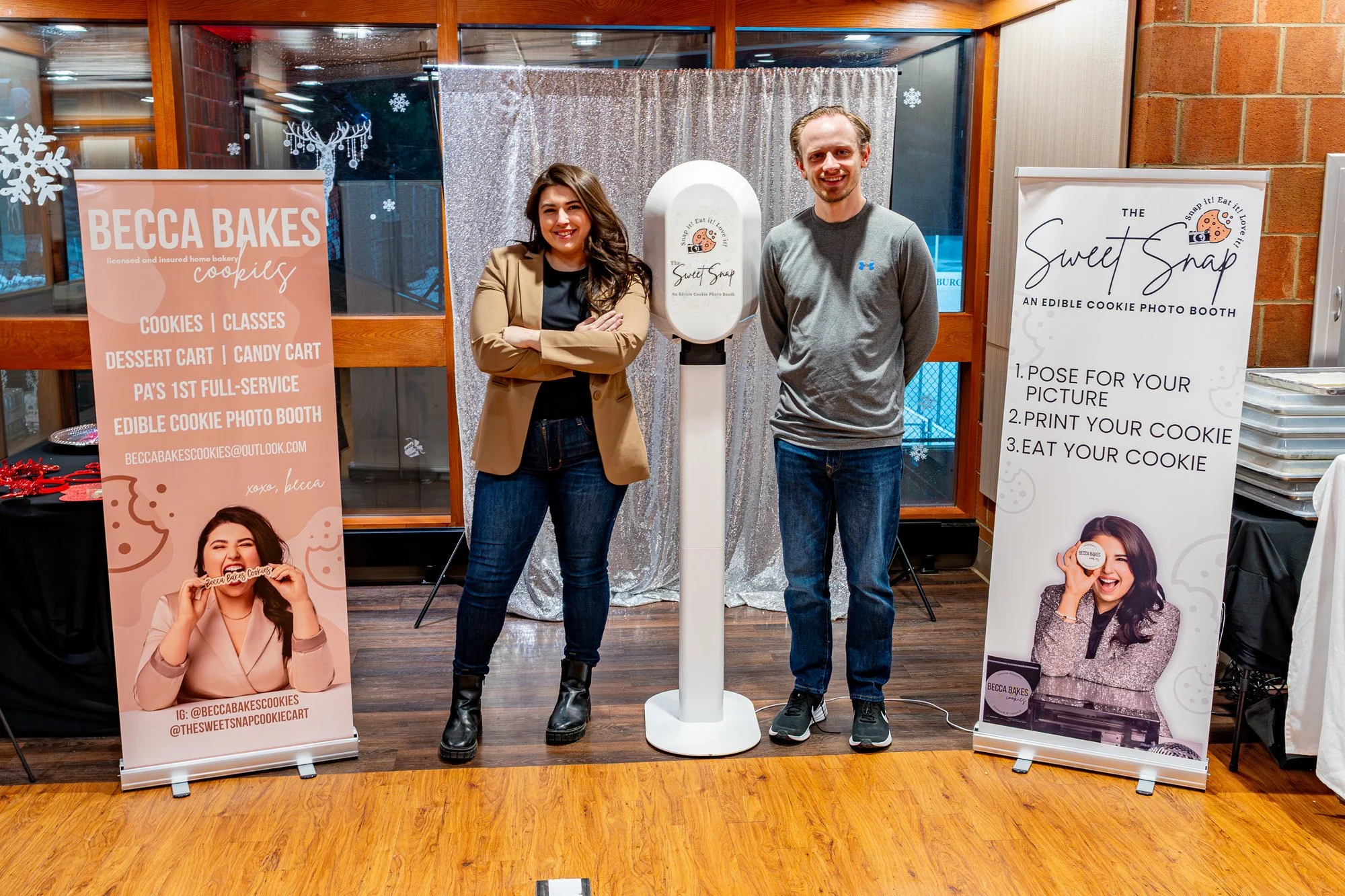 Two vendors stand beside an edible cookie photo booth display inside an indoor event space, framed by branded banners explaining the Sweet Snap cookie photo experience.
