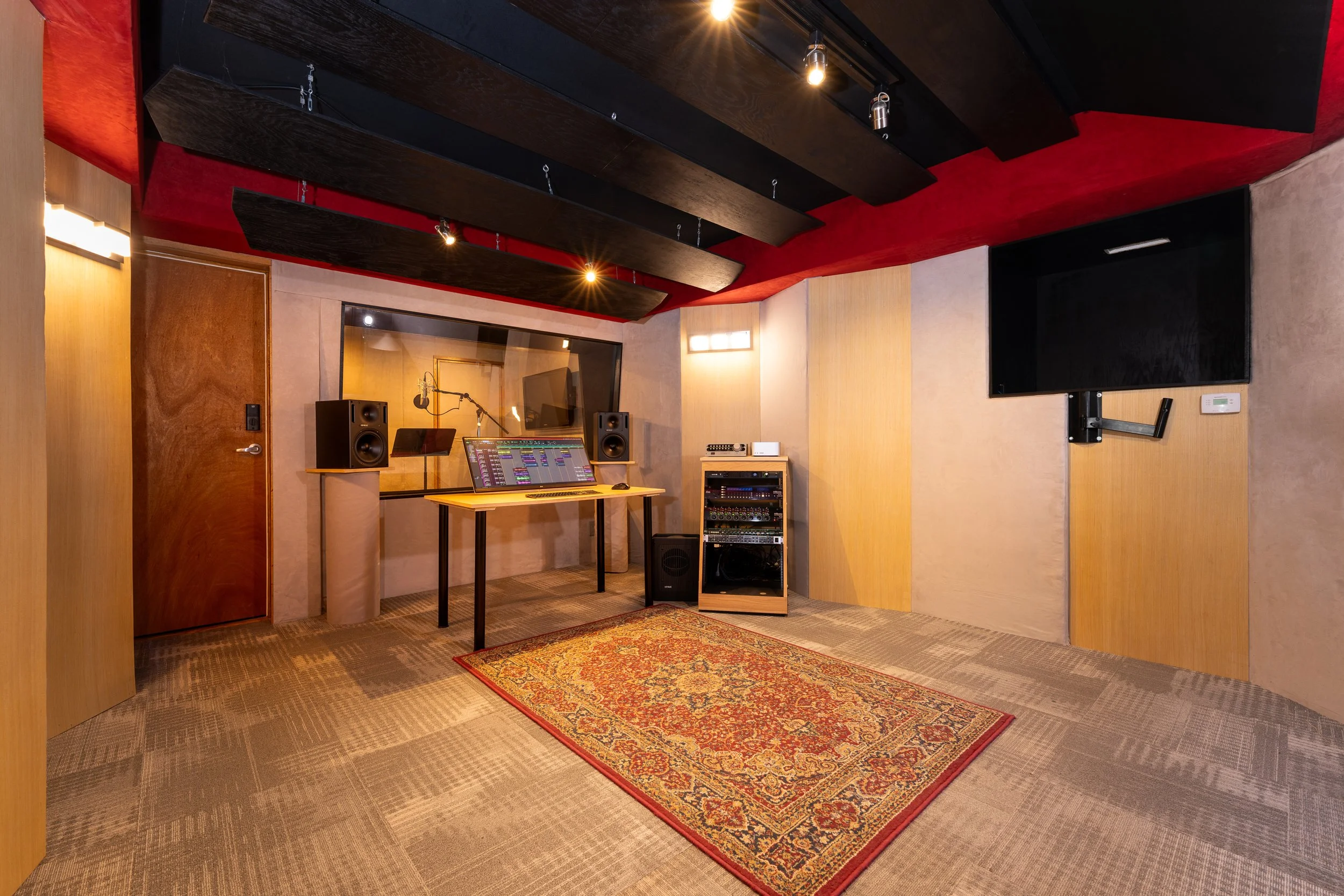 Recording studio control room with acoustic wall panels, ceiling baffles, a standing desk with computer and studio monitors, an equipment rack, and a patterned rug beneath warm overhead lighting.