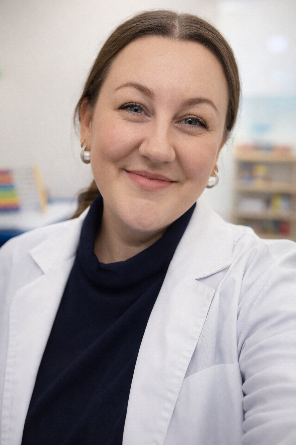 A smiling woman with light skin, brown hair, and wearing a white lab coat, taken in a professional setting like a medical office or laboratory.