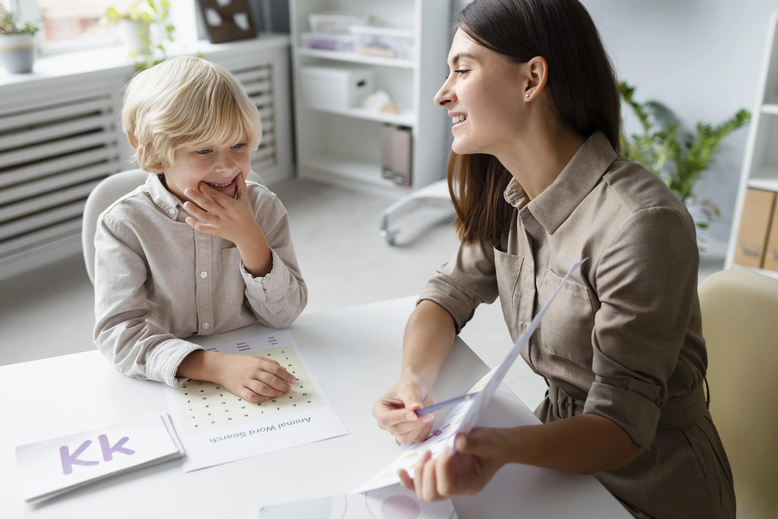 A young boy and a woman sitting at a table in a brightly lit room, engaging in an educational activity with flashcards and an artifical word search. The boy is smiling and touching his chin, while the woman is smiling and holding a booklet, possibly a teaching aid.