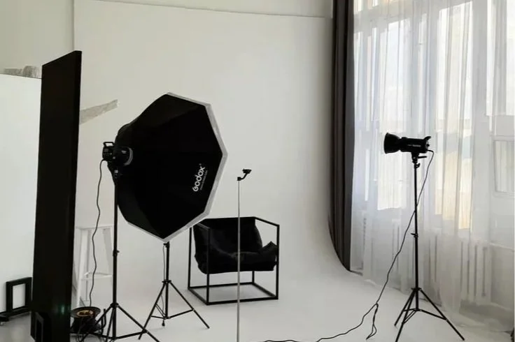 Photography studio setup with a backdrop, tripod, softbox light, camera on a stand, and a chair against a white wall with curtains.