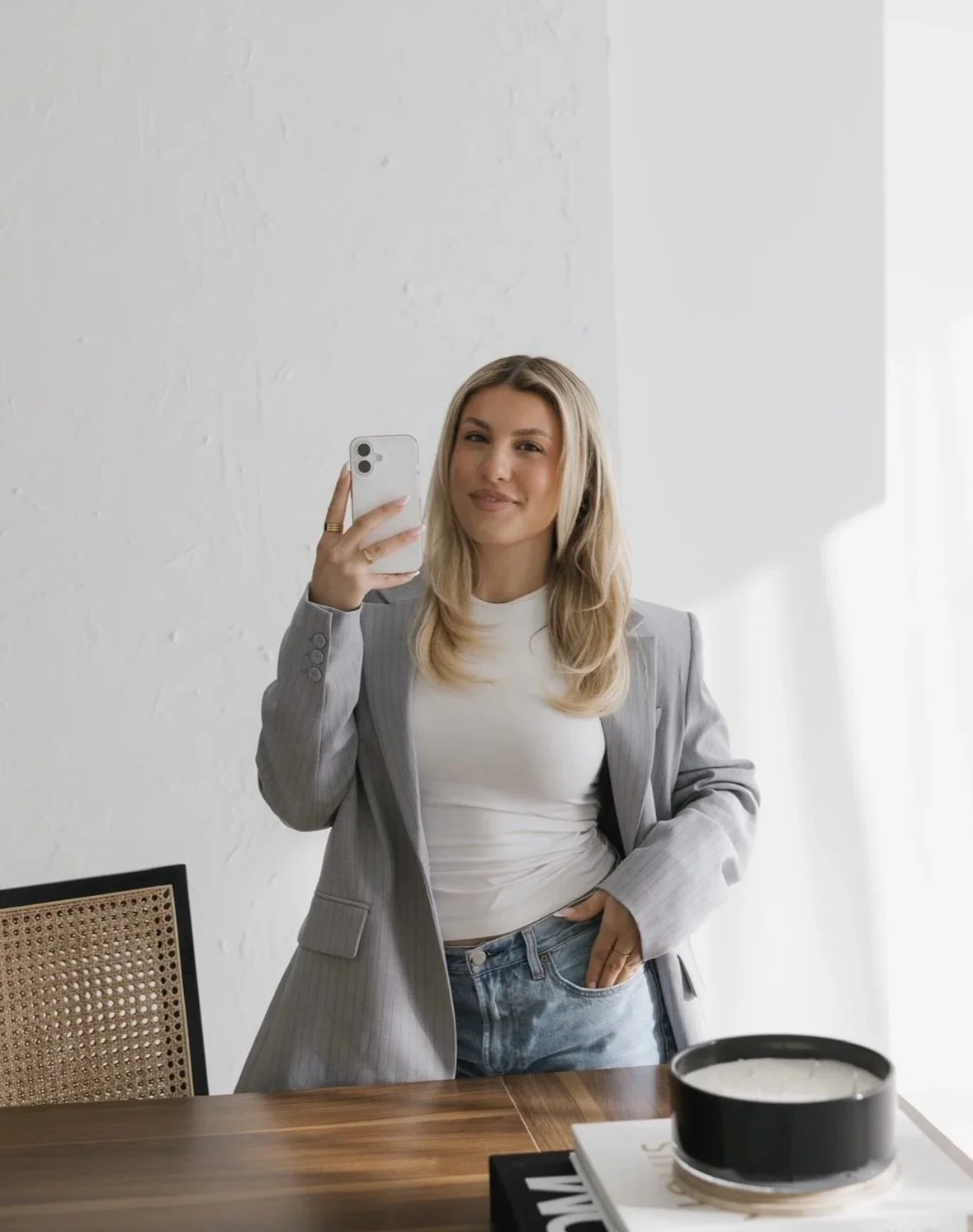 A woman with blonde hair taking a selfie in front of a mirror, wearing a gray blazer over a white shirt, standing in a minimalist room with a wooden table.