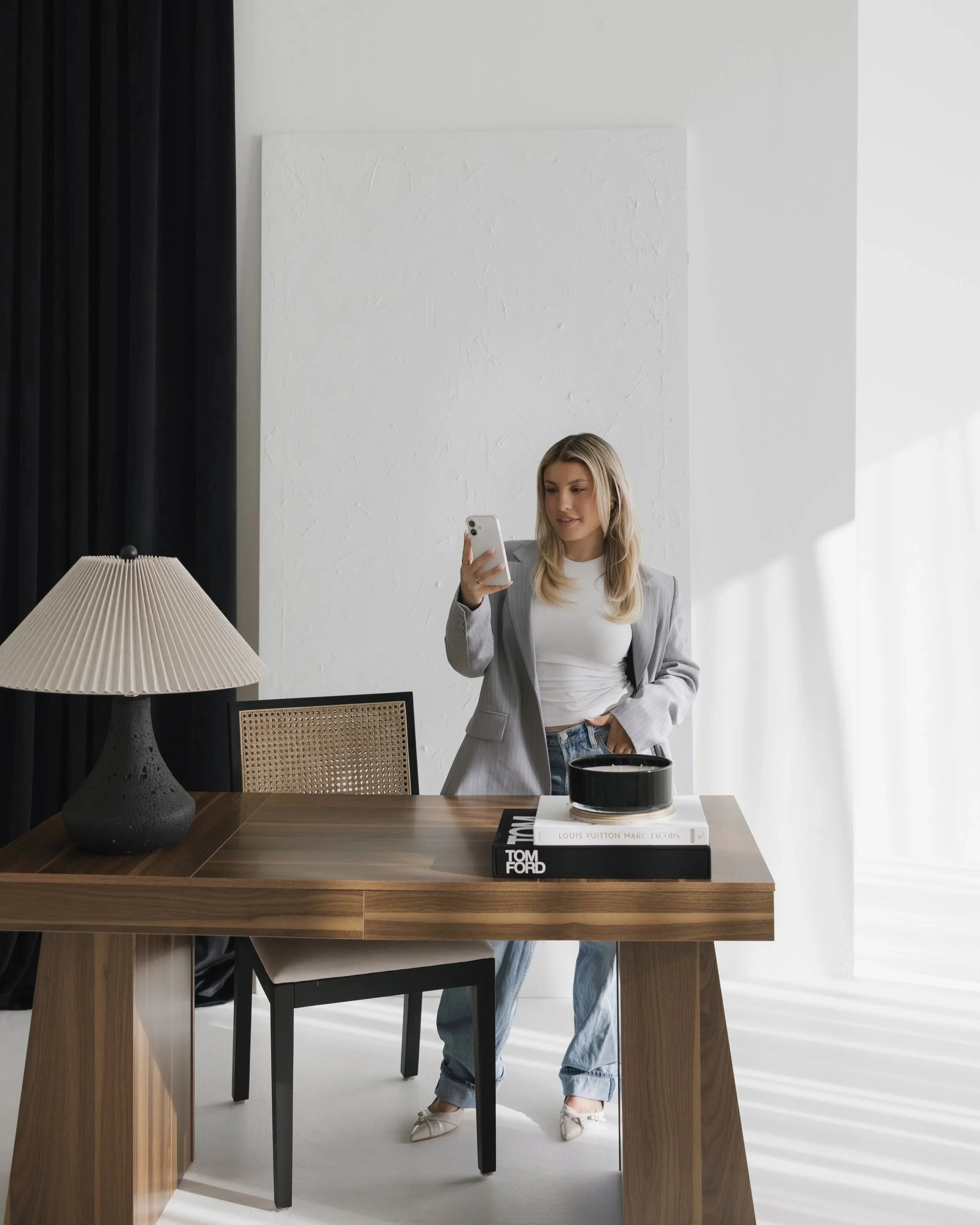 A woman in a gray blazer and jeans holding a smartphone stands behind a wooden table in a modern, minimalistic room. The table has a large stack of books titled Louis Vuitton, Marc Jacobs, and Tom Ford, with a black bowl on top. A black curtain is on the left, and a black table lamp with a pleated beige lampshade is in the foreground. The background features a white textured wall and a large window with sheer white curtains.