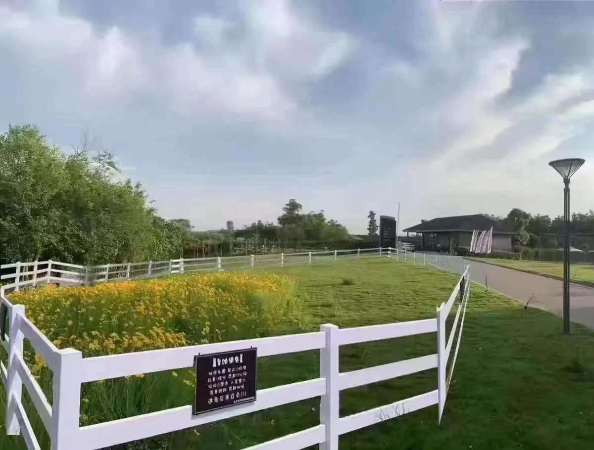 A fenced garden with yellow flowers, green grass, a path, and a building in the background, under a partly cloudy sky.