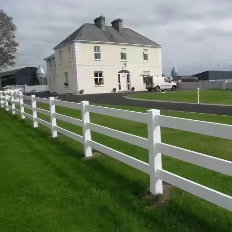 A large cream-colored house with a dark roof, white window frames, and a small front door. There is a white picket fence surrounding a well-maintained green lawn, a driveway with a parked white van, and nearby outbuildings or structures under a cloud