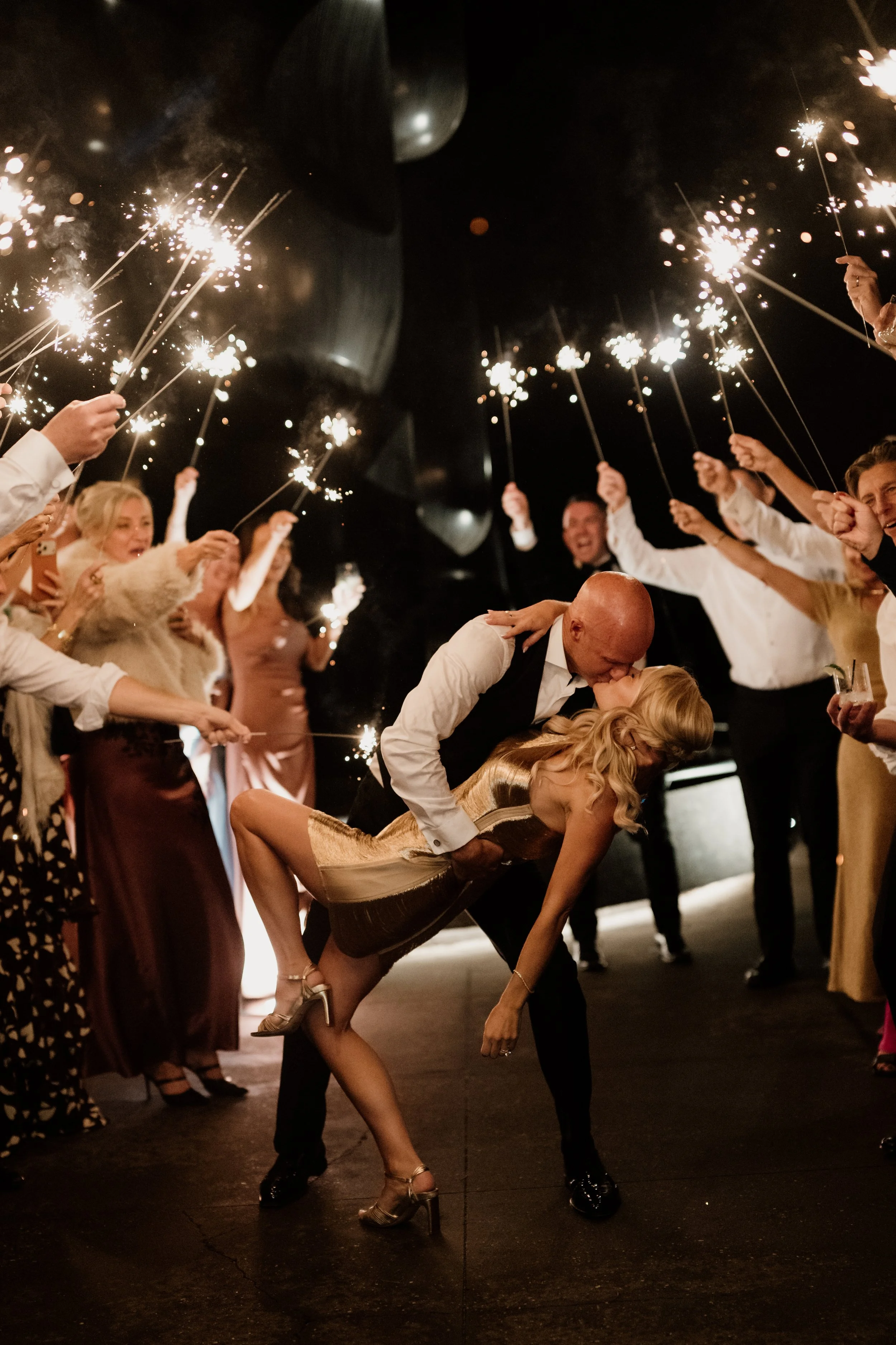 A man and woman kissing as they dance, surrounded by guests holding sparklers during a celebration event.