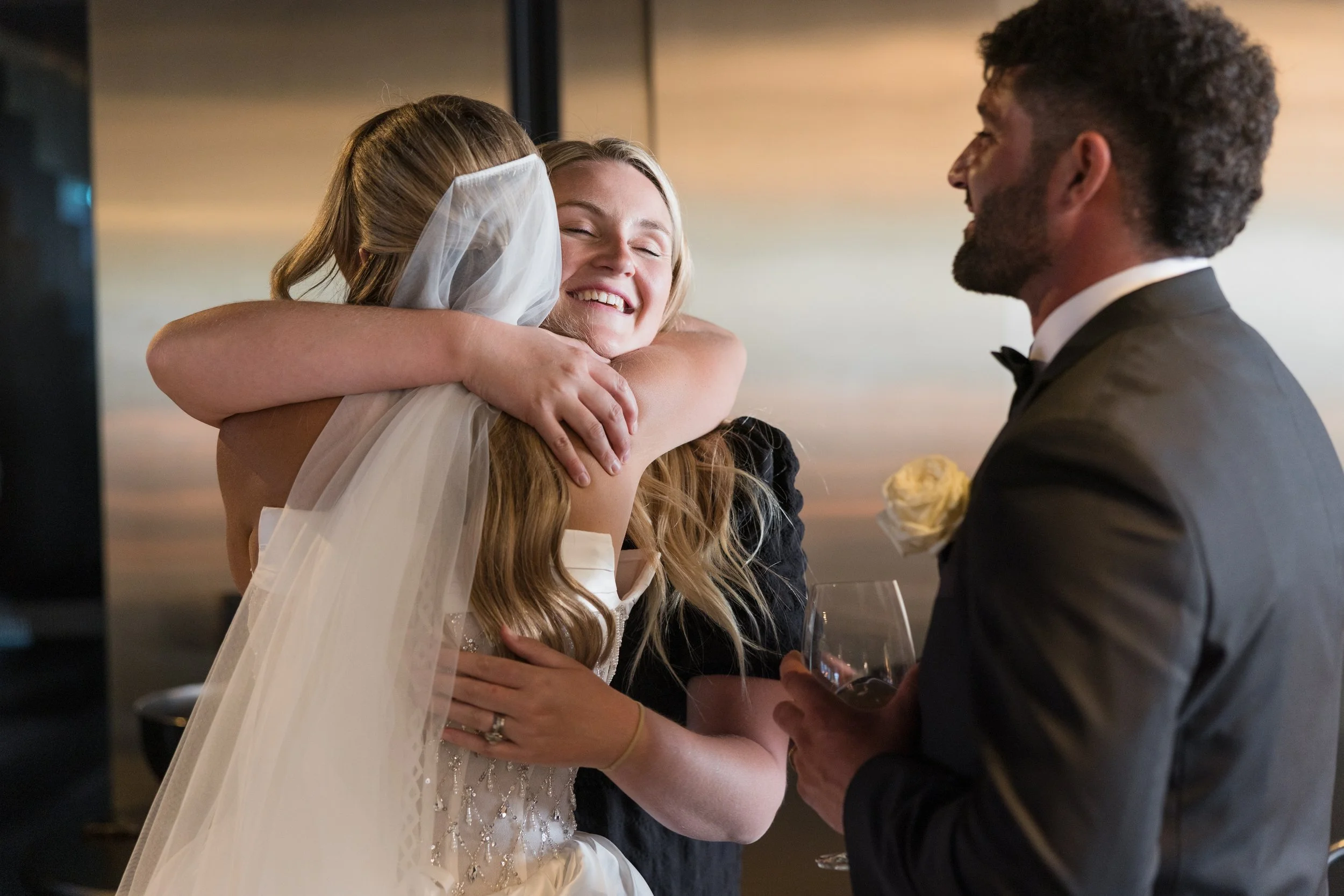 A bride and a woman hugging, and a groom holding a wine glass, during a wedding celebration.