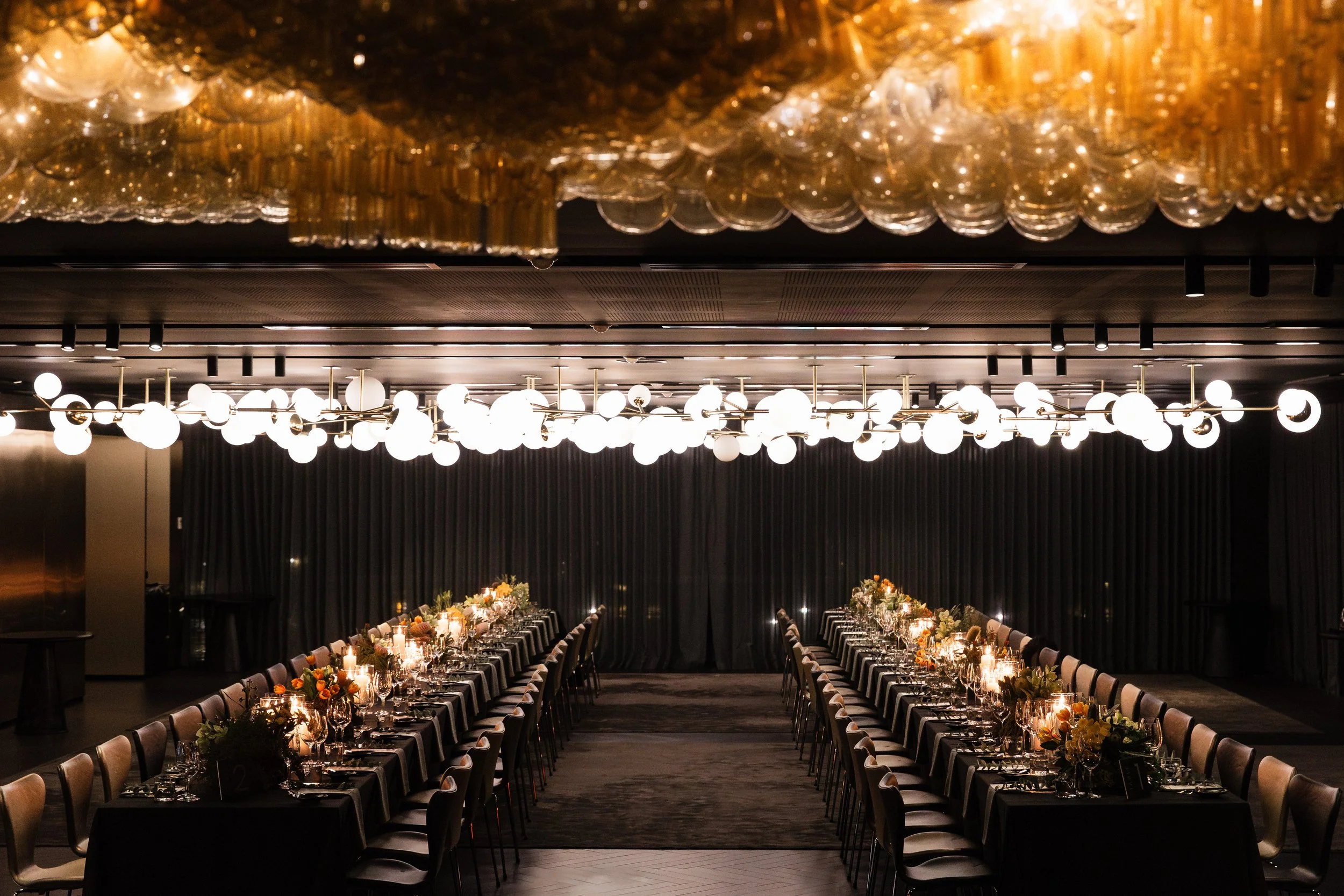 A banquet hall with two long tables set for a formal dinner, decorated with floral centerpieces and candles, with modern lighting fixtures overhead, and dark curtains in the background.
