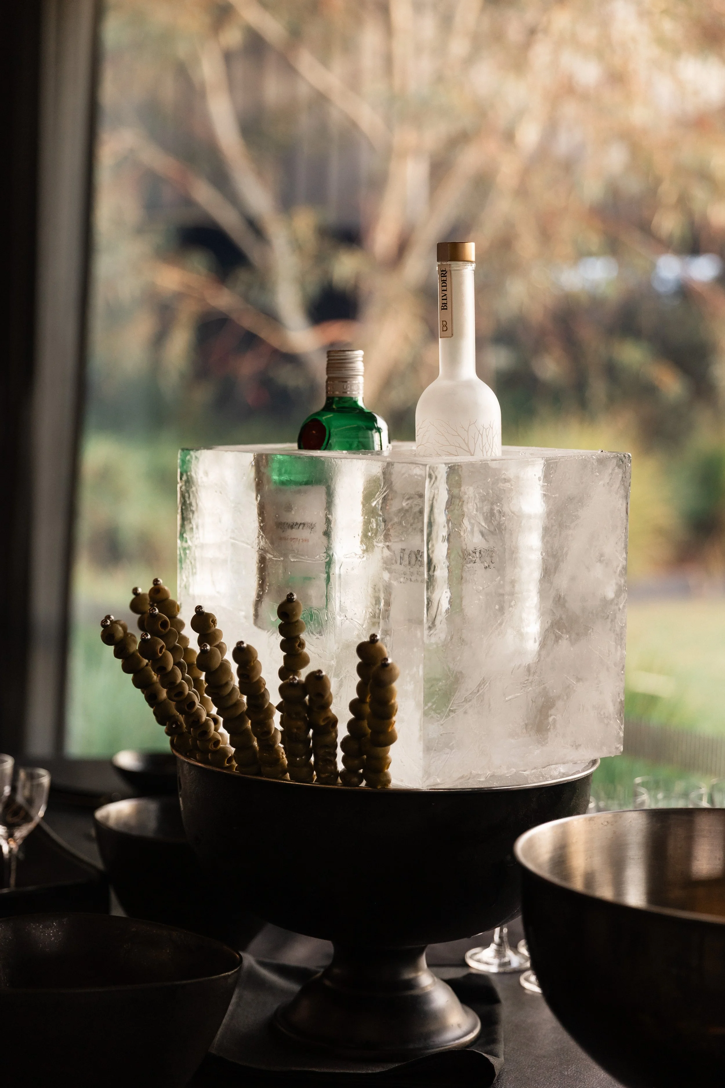 Bottles placed on an ice block with skewers of olives in bowls, set indoors with a window showing trees outside.