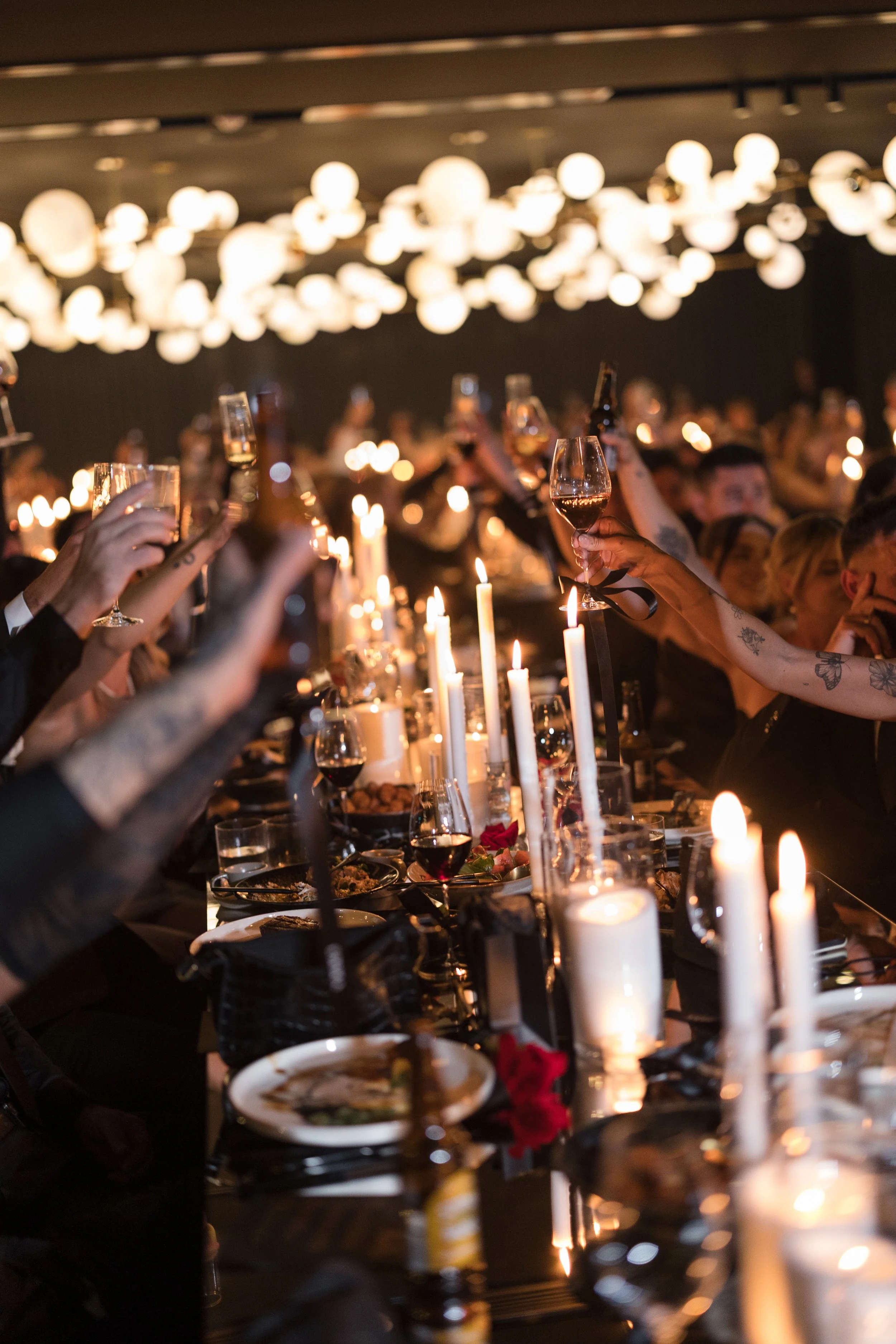 People raising glasses at a dinner party with candles on the table and modern light fixtures hanging from the ceiling.