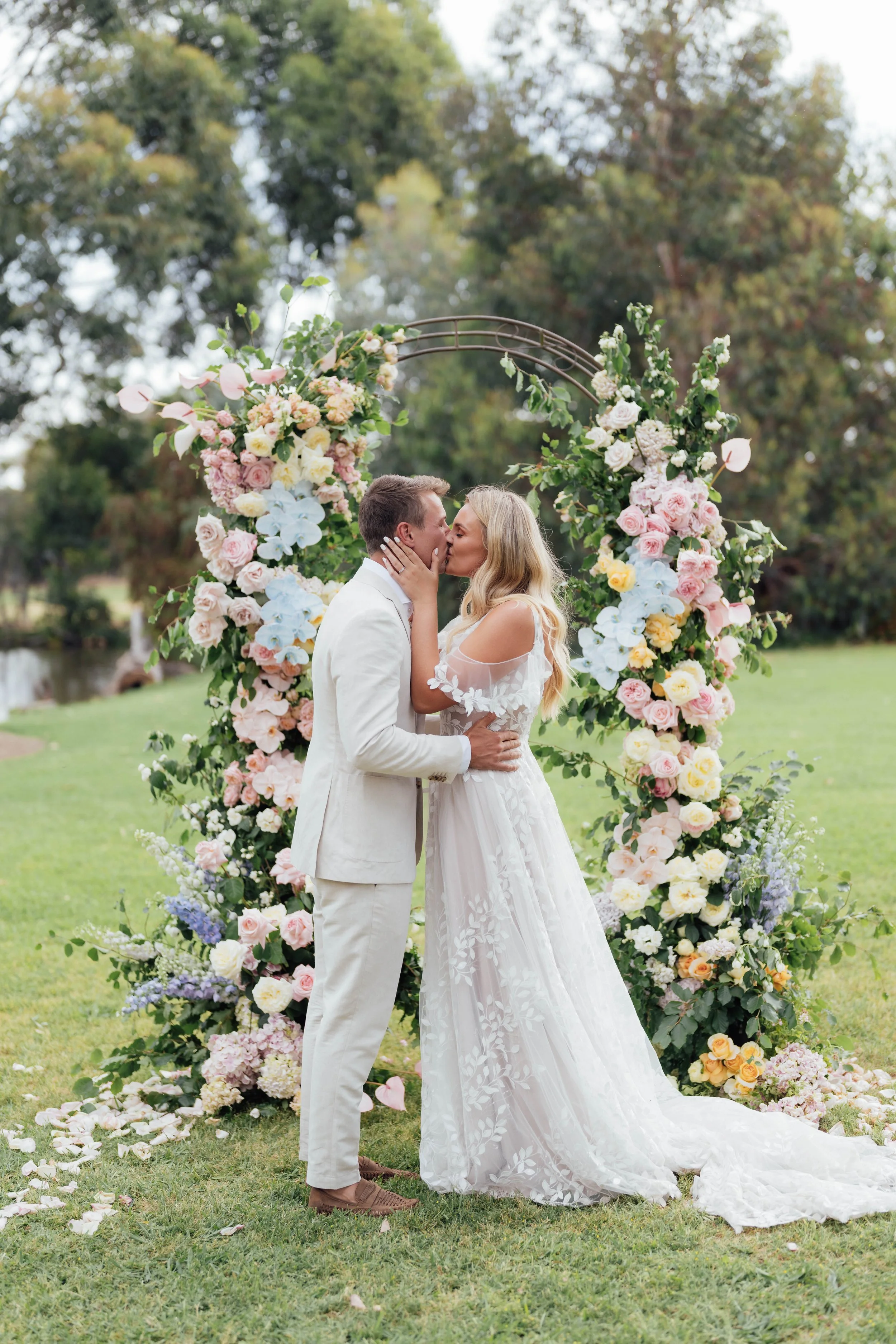 A bride and groom share a kiss in front of a floral wedding arch outdoors.
