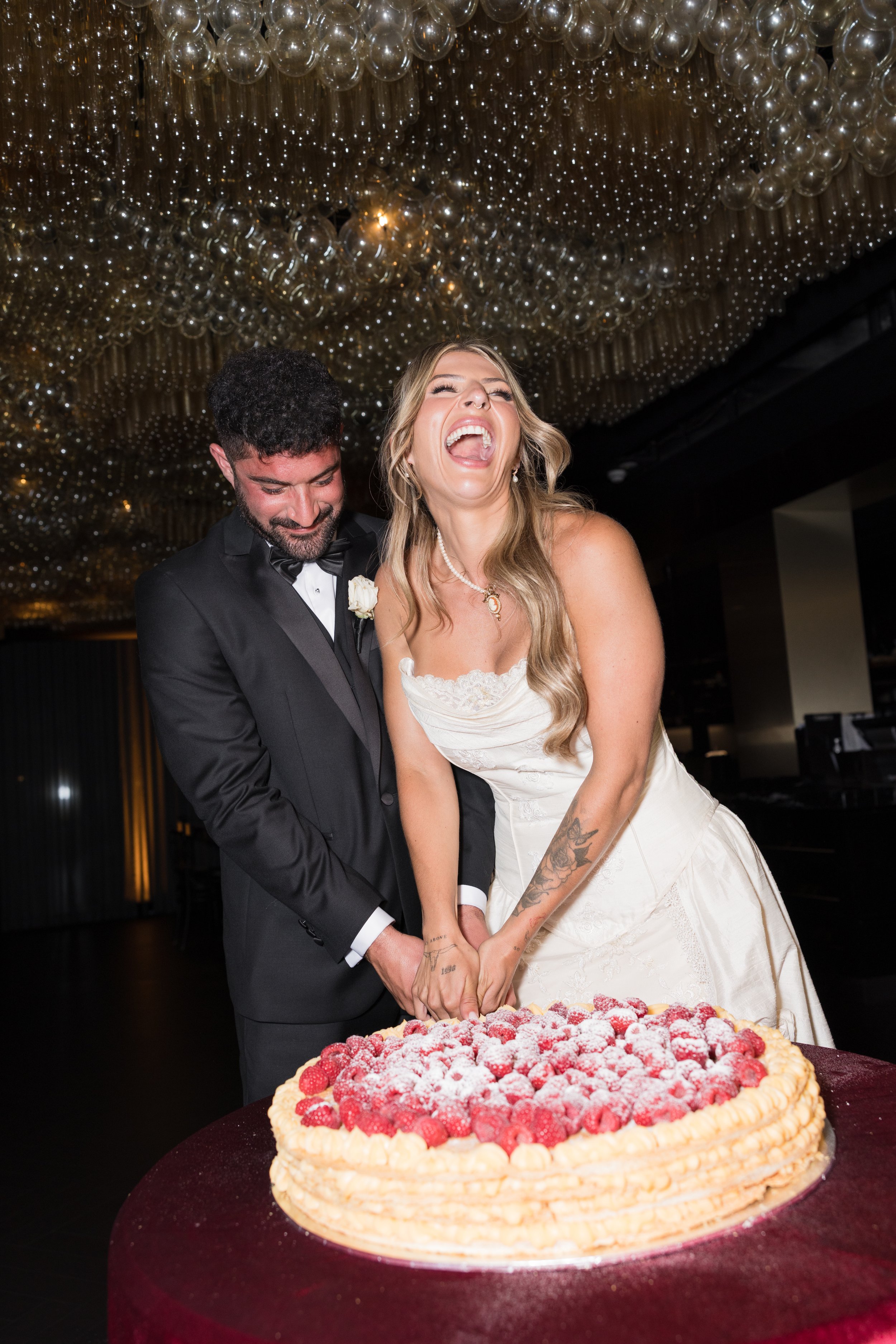 Bride and groom cut a wedding cake together at their wedding reception, both smiling and laughing.