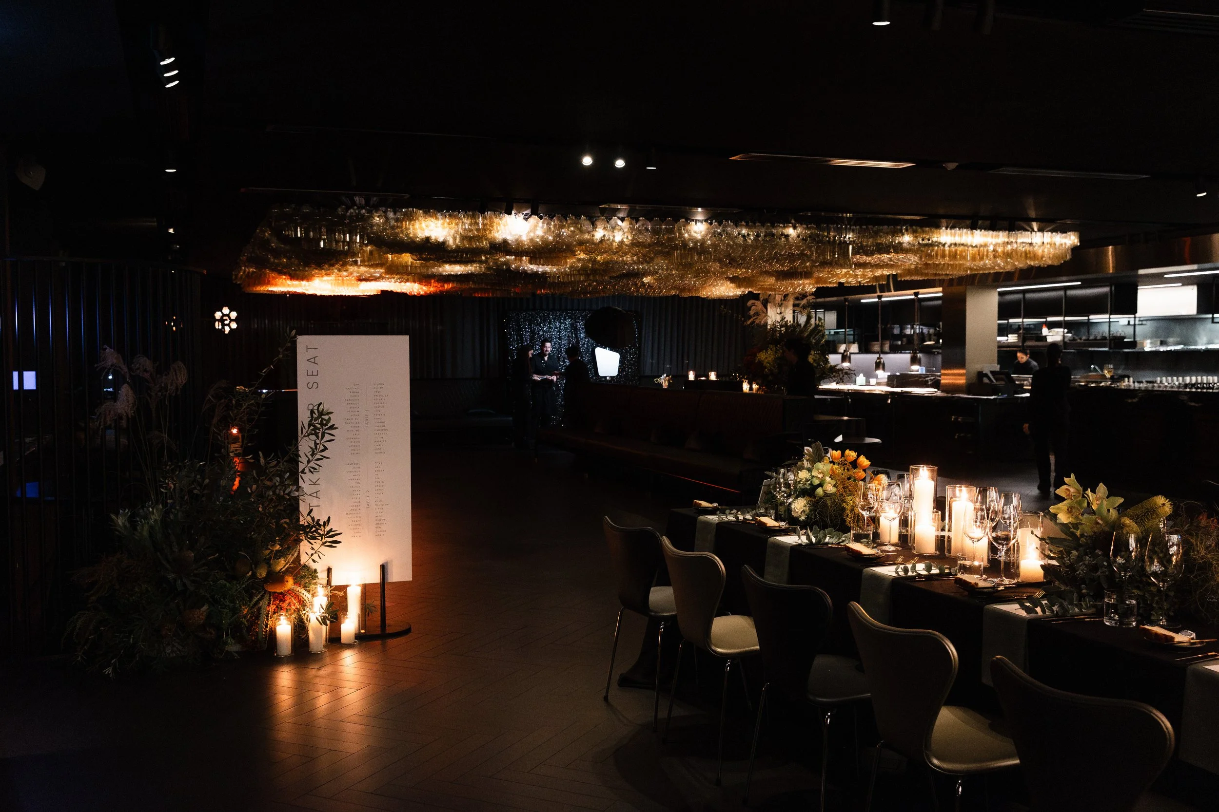 Dimly lit restaurant with a long table set with candles, flowers, and glasses, and a bar in the background.