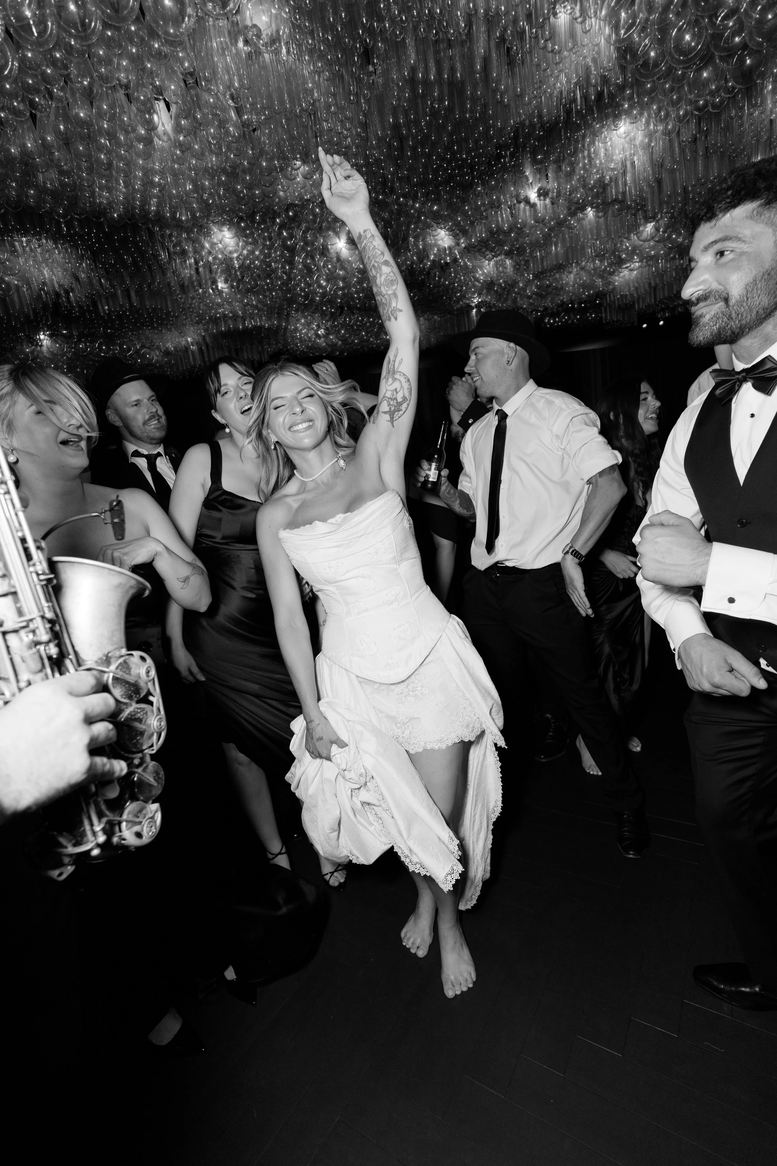 People dancing and celebrating at a party, woman in a white dress raising her arm, others surrounding her in formal attire, with a decorative ceiling overhead.