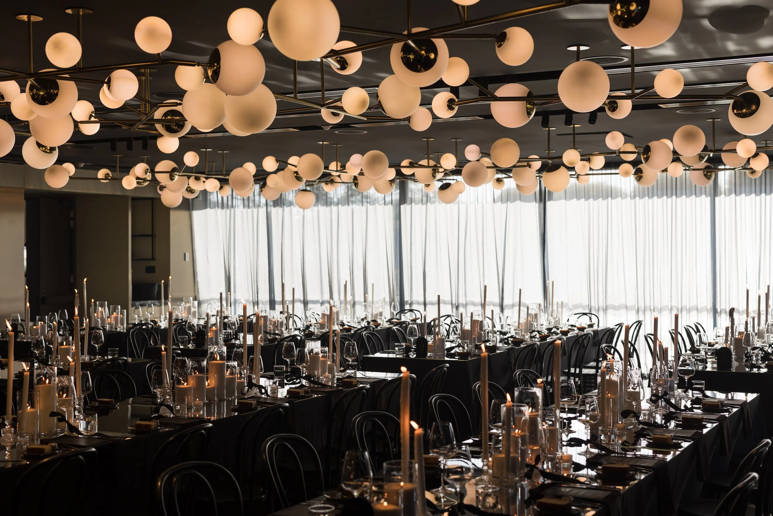 A banquet hall decorated with a large number of spherical light fixtures on the ceiling and tables set with candles, glasses, and cutlery, with large windows and curtains in the background.