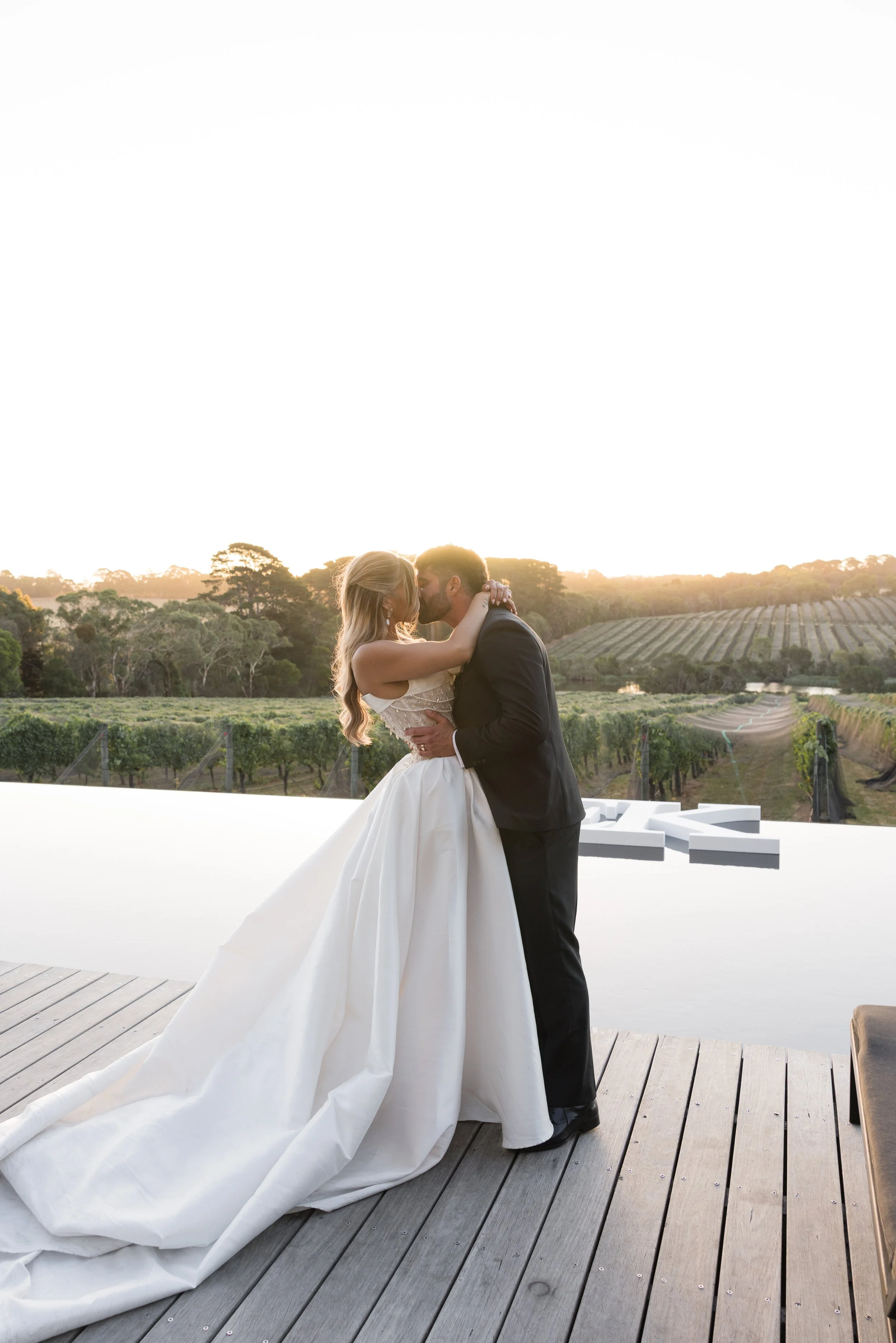 Jackalope wedding. Bride and groom kiss during sunset in vineyard setting with wooden deck and modern white art pool float in background.