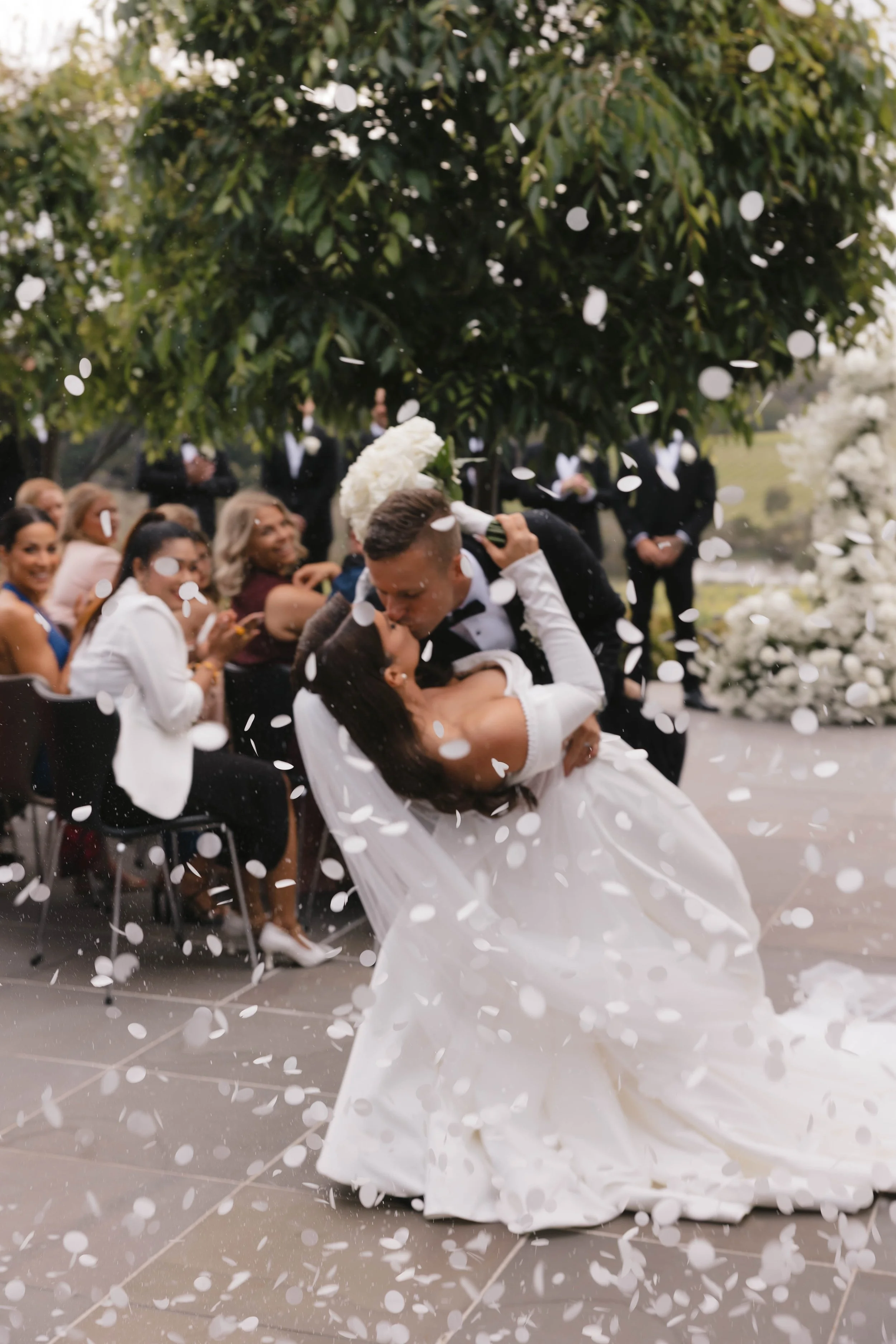 Couple kissing during wedding celebration with guests watching, white flower petals falling, outdoors with trees and floral decorations.