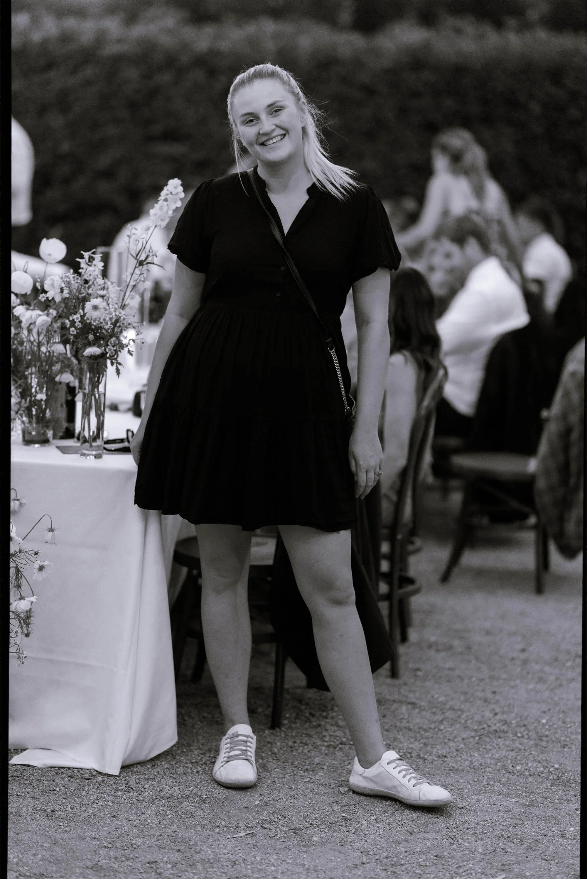 A young woman with blonde hair tied back, smiling, wearing a black dress and white sneakers, standing outdoors at a social gathering with a table decorated with flowers on her left and people sitting and standing in the background.
