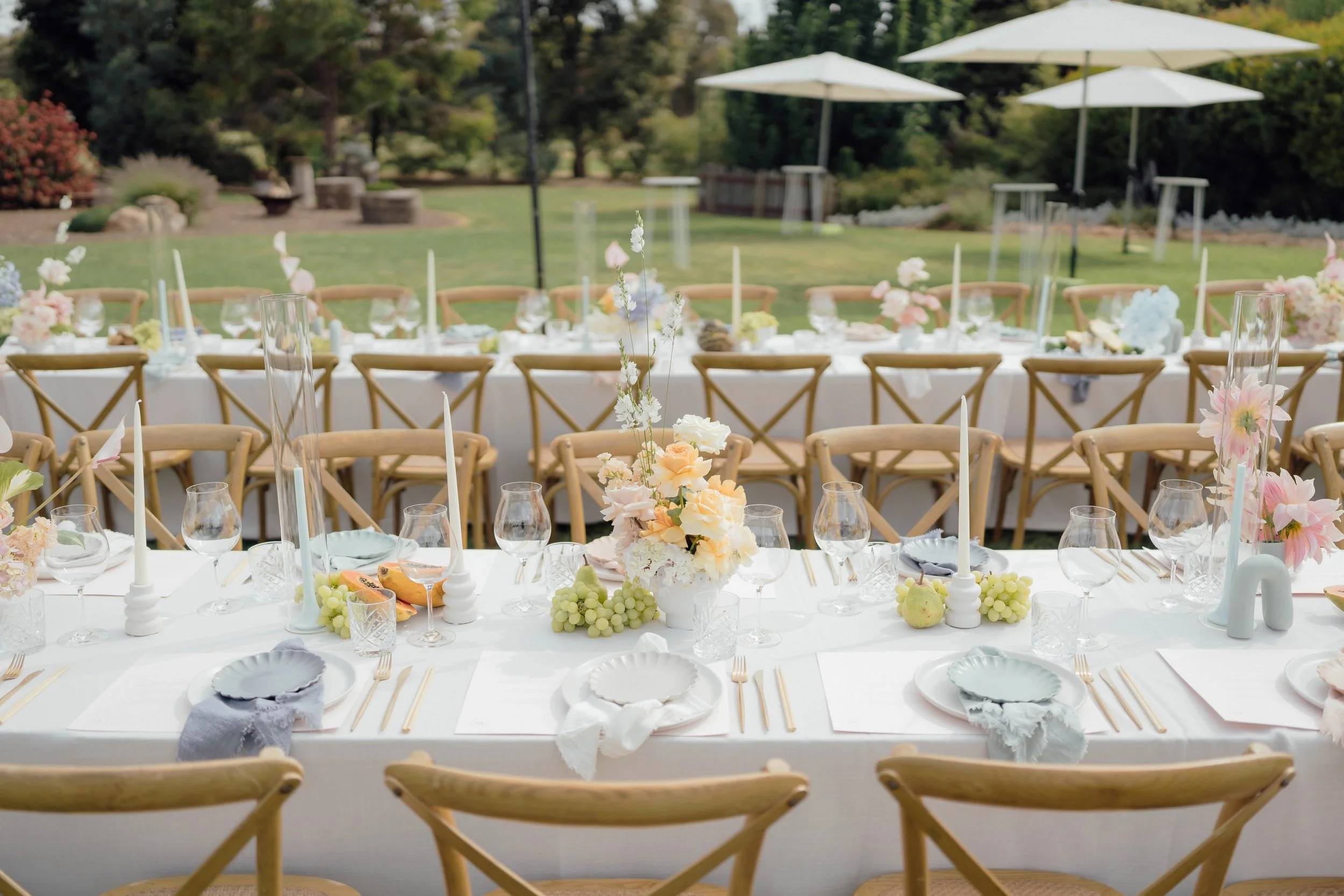 Elegant outdoor wedding reception table setup with white tablecloth, floral centerpieces, candles, glassware, and table settings, surrounded by wooden chairs, set on a lush green lawn with trees and umbrellas in the background.