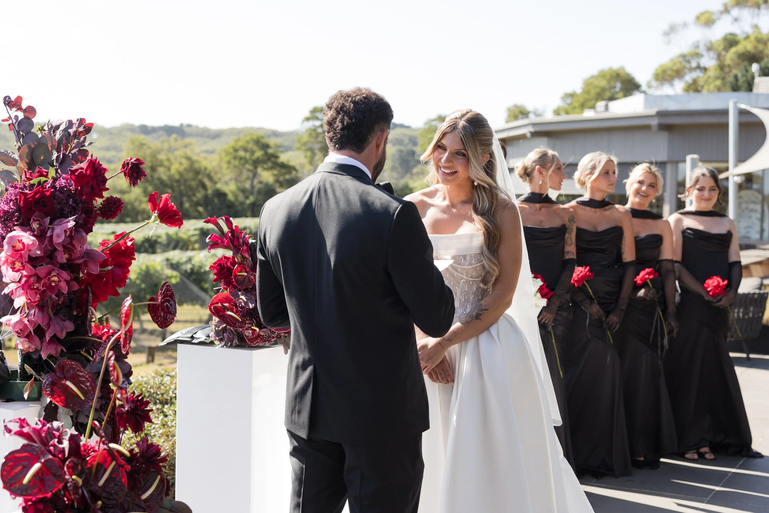 A wedding ceremony outdoors with a bride and groom exchanging vows, surrounded by bridesmaids in black dresses holding red flowers, and decorated with red and pink flowers in a natural setting.