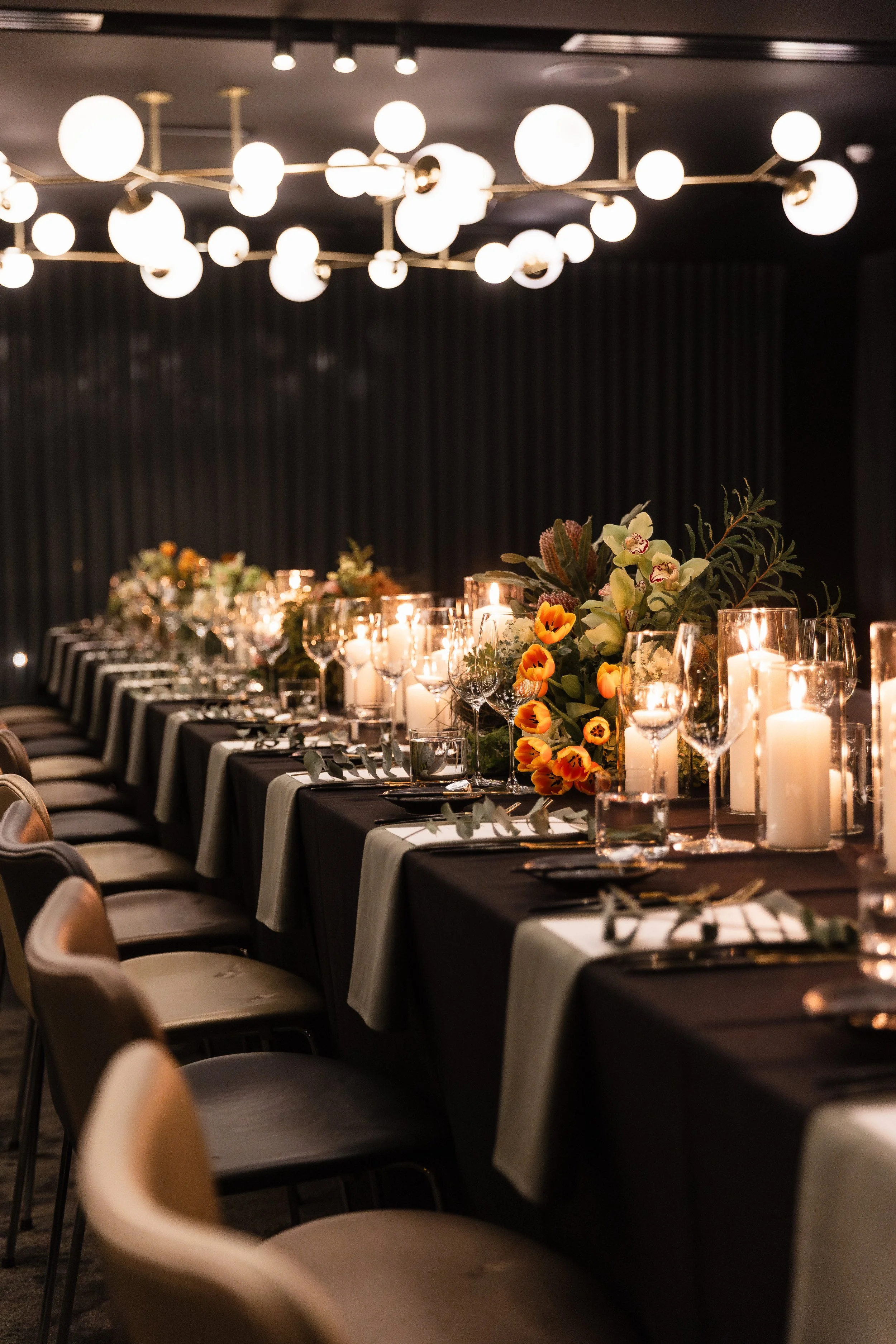 A long banquet table decorated with floral centerpieces and lit candles in a dimly lit elegant dining room.