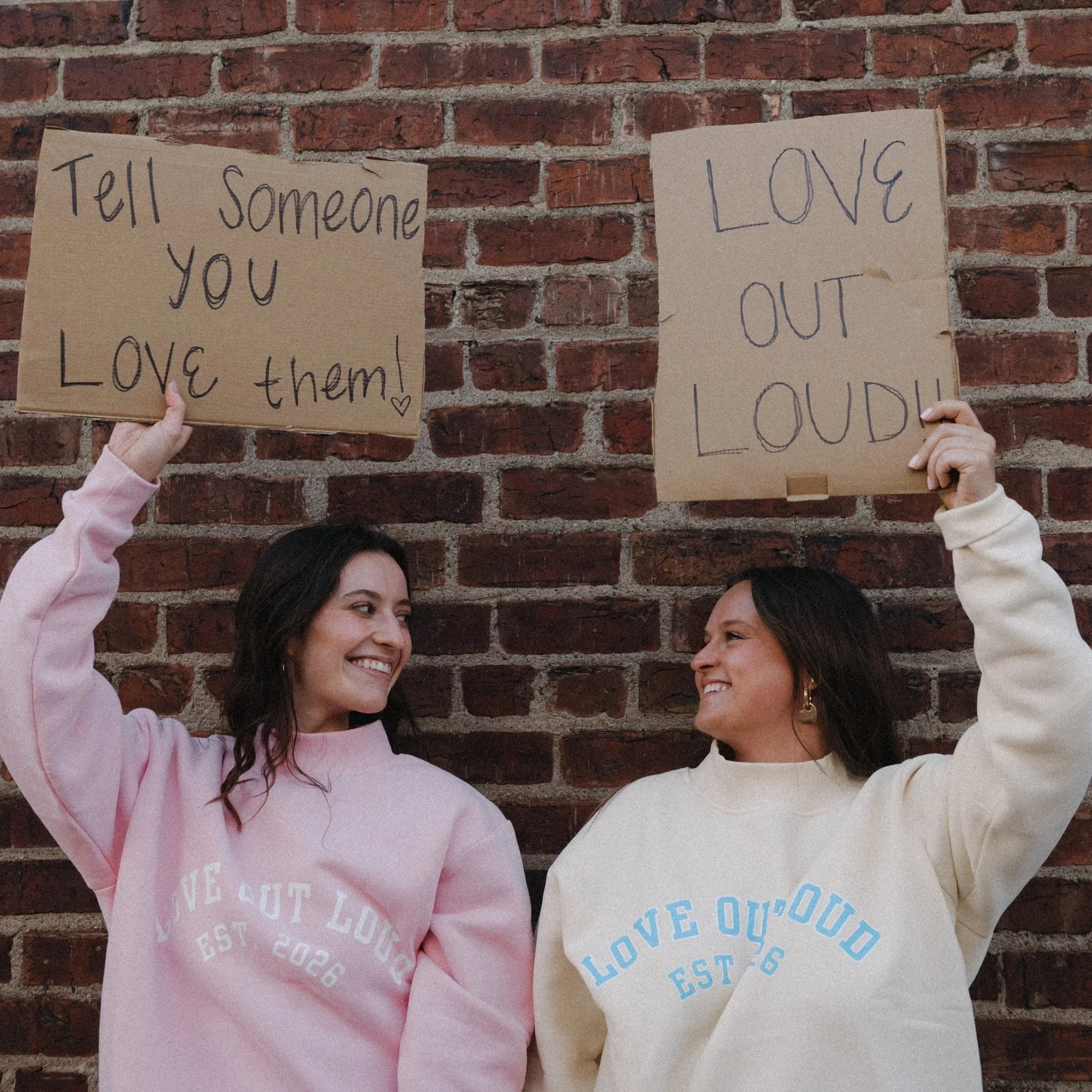 Four people holding handmade signs in front of a brick wall, with two women visible smiling at each other.