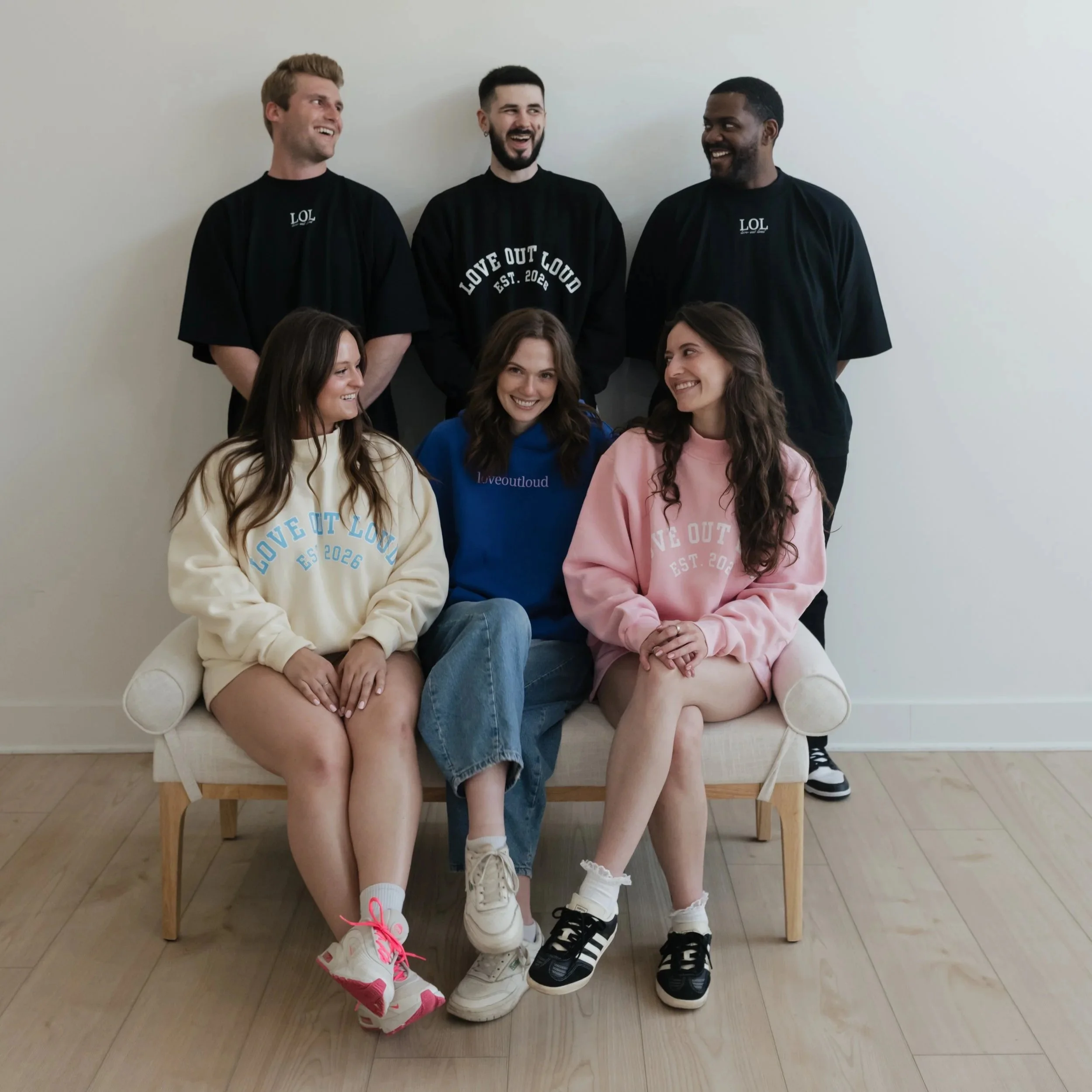 Group of five young people sitting on a wooden bench against a white wall, smiling and engaging in conversation. Four are standing behind, three women and one man, and two women are seated in front. They're dressed casually in colorful sweatshirts an
