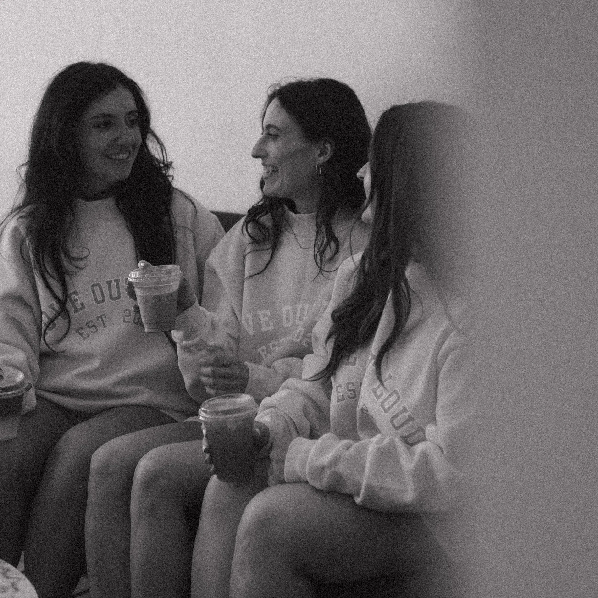 Three women closely talking and smiling, wearing casual clothing, one holding a drink, in black and white.