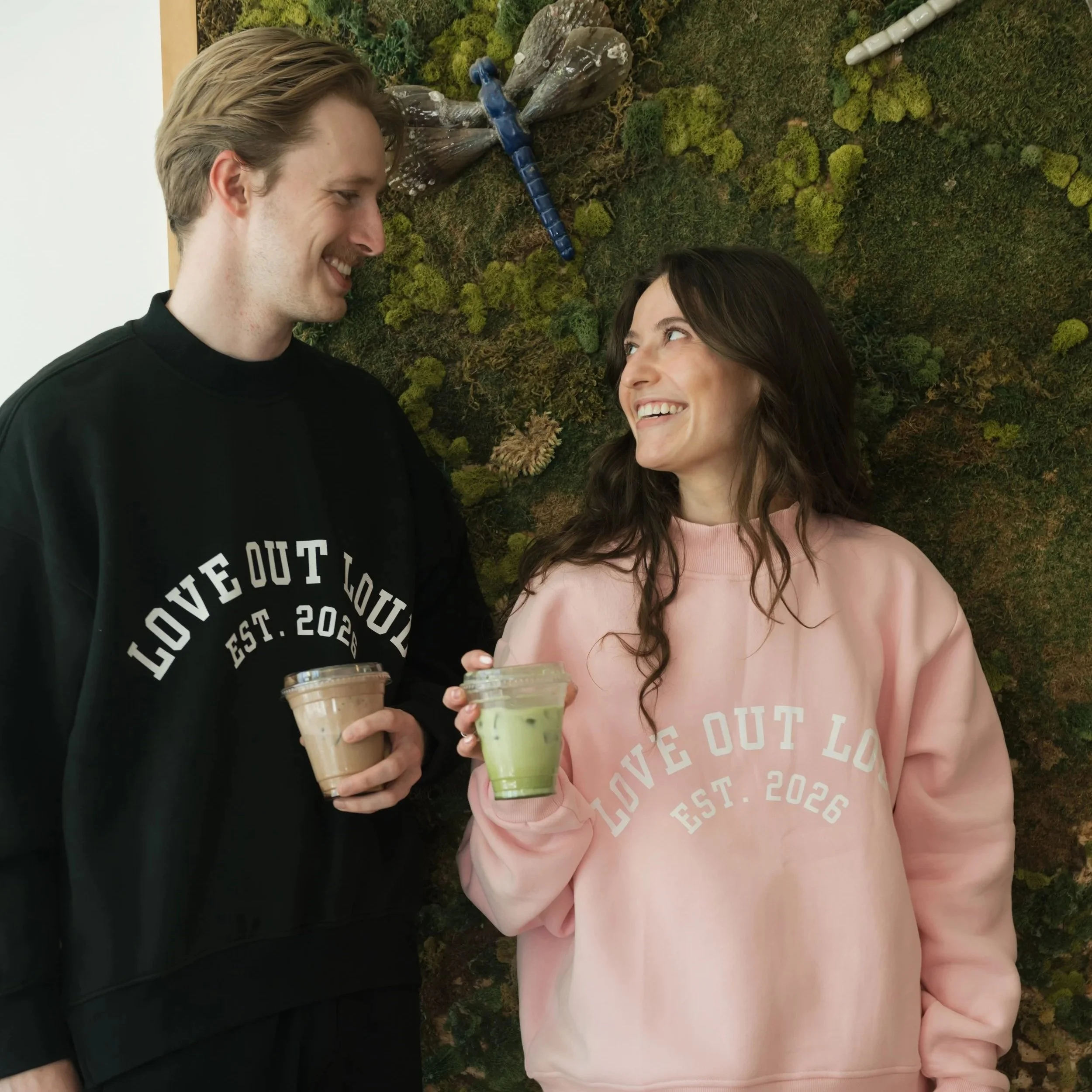 A man and woman smiling at each other while holding coffee drinks, standing in front of a moss-covered wall